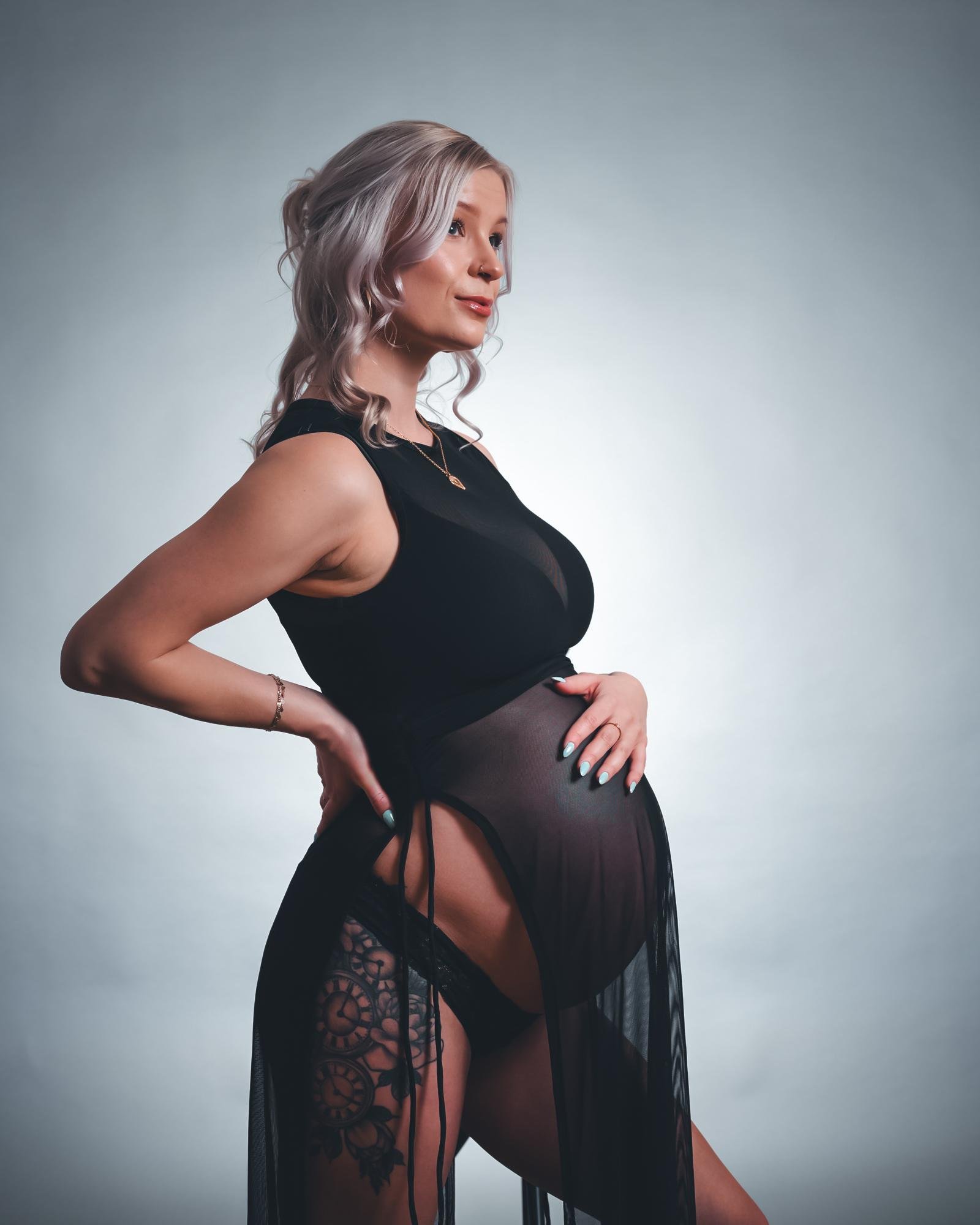 Pregnant woman with light blonde hair in a black sleeveless dress, posing in profile against a light gray background. She is holding her belly with both hands and wearing sheer black fabric around her waist revealing intricate tattoos on her thigh.