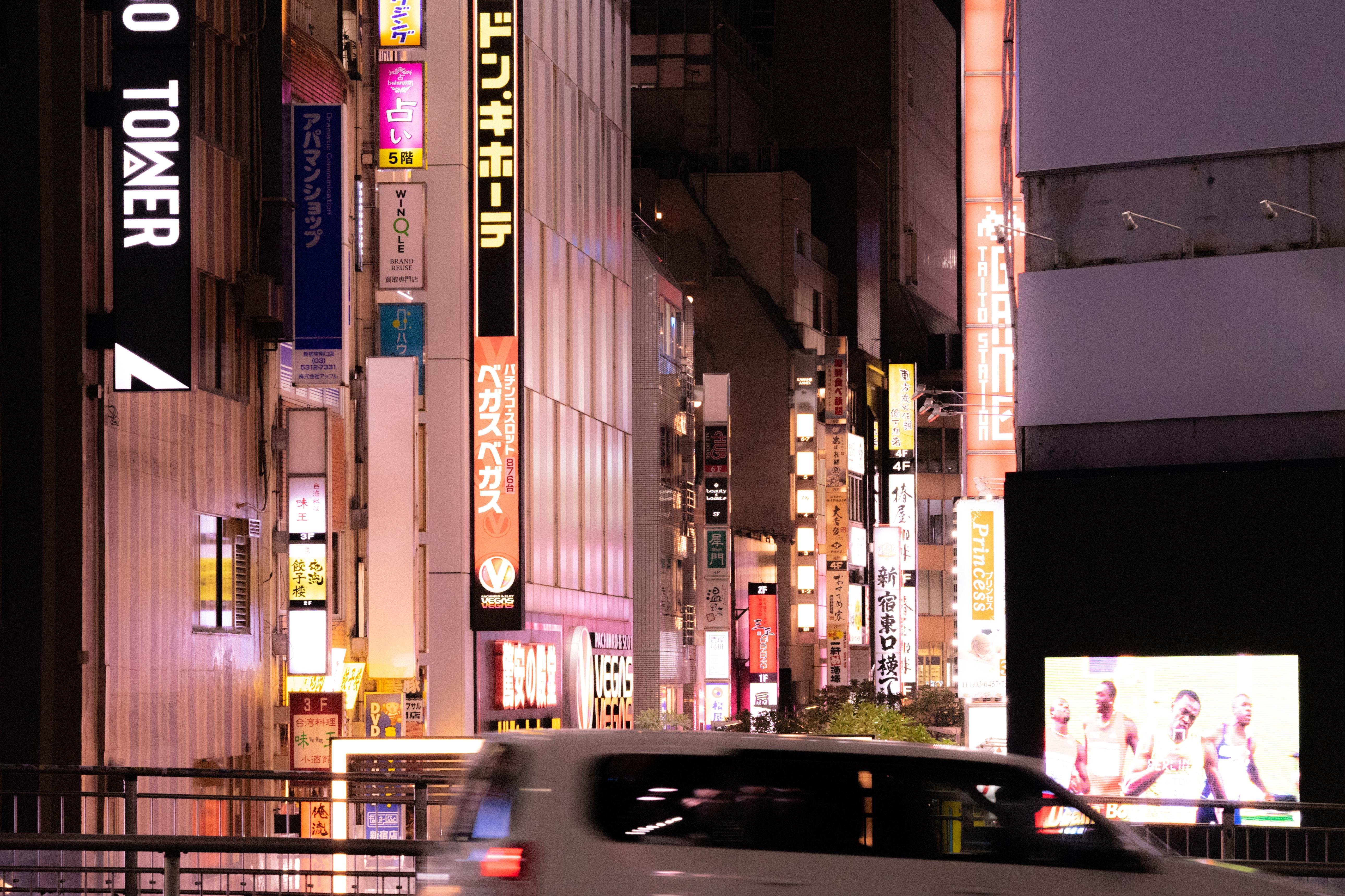 City street at night with numerous brightly lit vertical signs in Japanese. Buildings line both sides of the narrow street, appearing densely packed. A white SUV is blurred in the foreground, indicating motion. An illuminated digital billboard on the right displays people in colorful clothing.