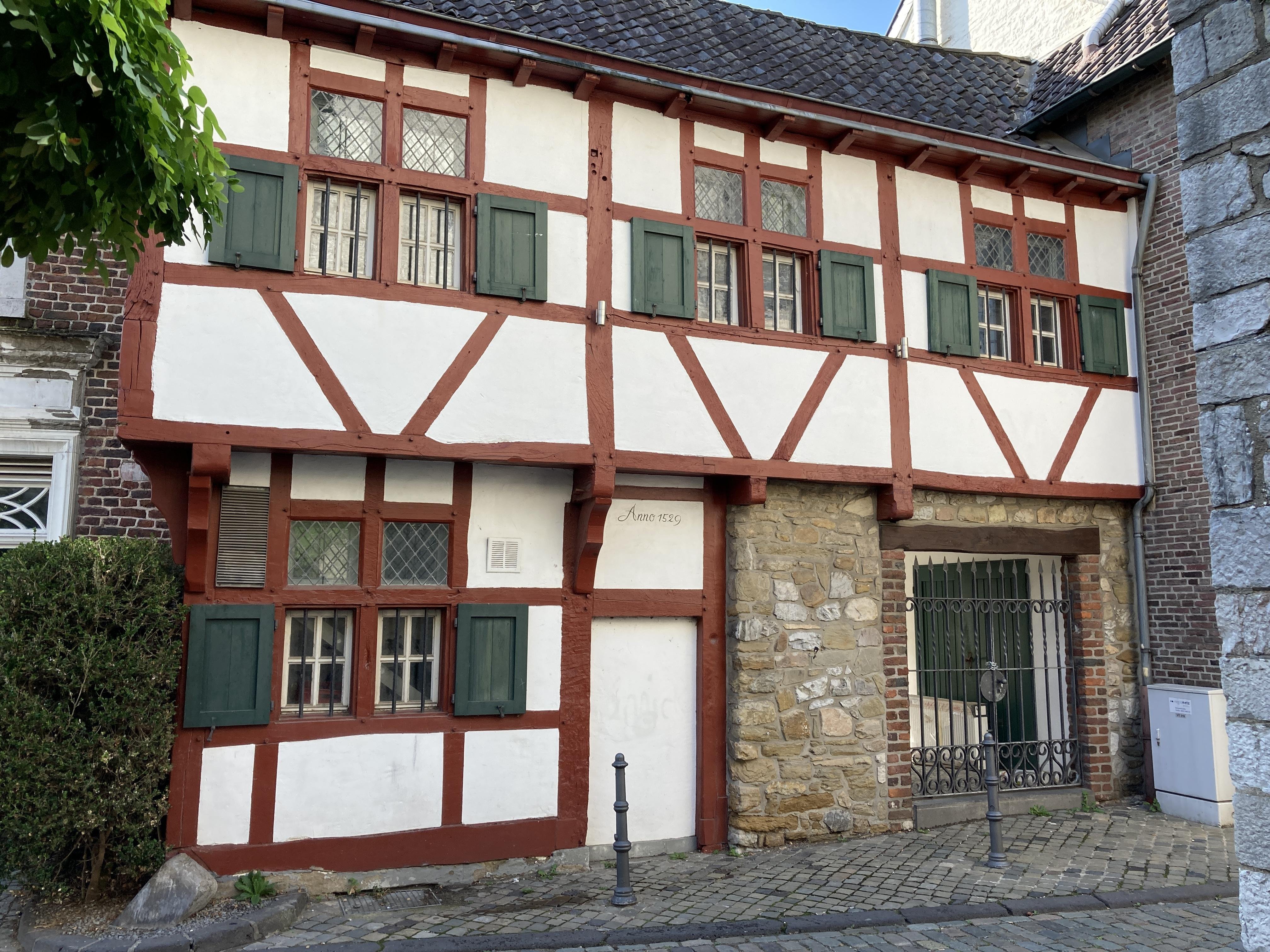 Das Bild zeigt ein Fachwerkhaus mit roten Holzbalken und weißen Wänden. Die Fenster sind mit grünen Fensterläden versehen. An der Fassade ist die Jahreszahl „Anno 1529“ zu lesen. Ein Teil des Gebäudes besteht aus Natursteinmauerwerk. Vor dem Haus befindet sich ein schmiedeeisernes Tor und Kopfsteinpflaster. Das Haus wirkt historisch und gut erhalten.