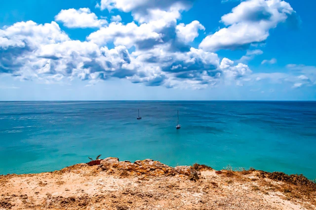 Cliffside view of a vast ocean with two sailboats in the distance. Clear turquoise water stretches to the horizon under a partly cloudy blue sky. The foreground features rocky, sandy terrain with sparse vegetation.