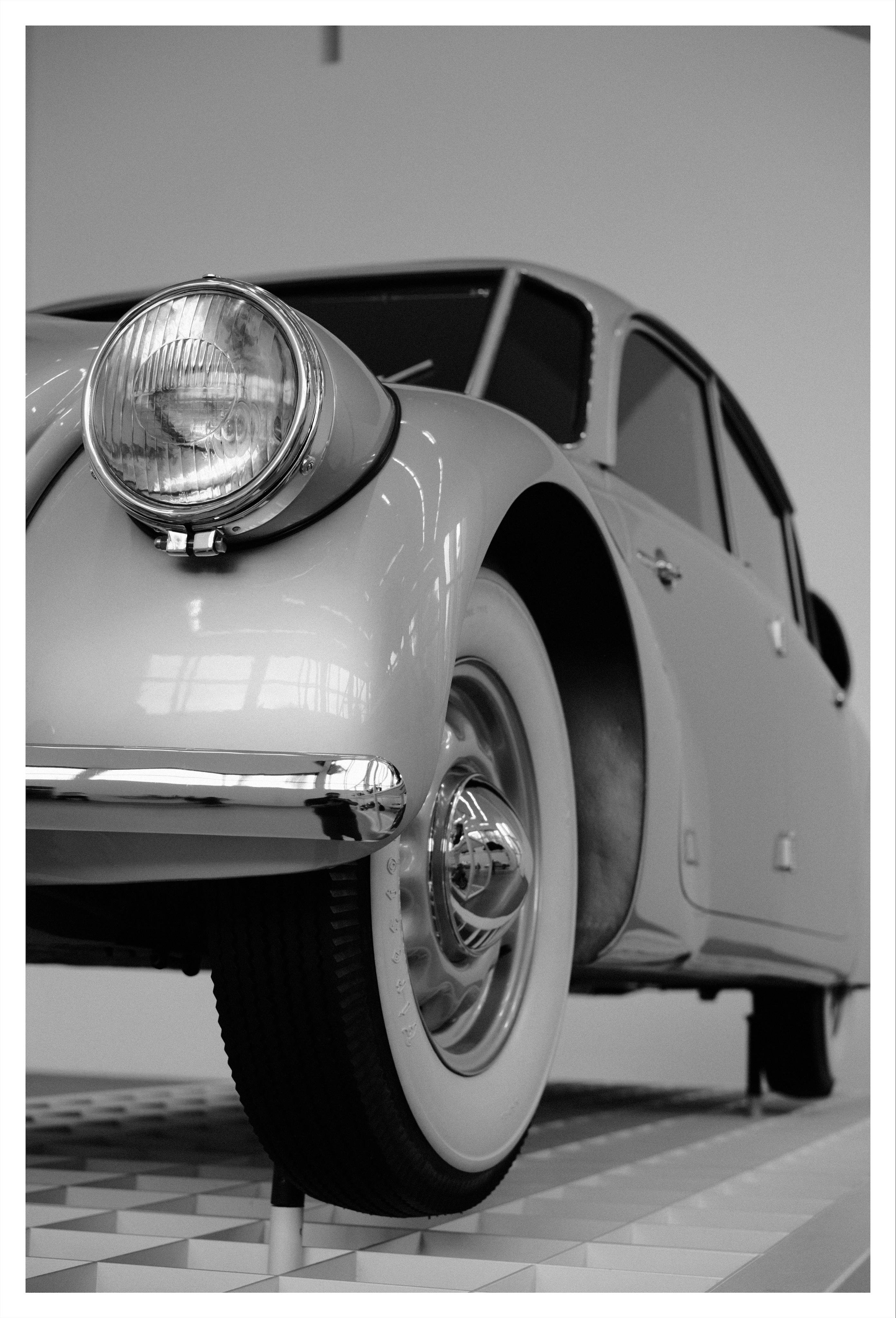 Low-angle black-and-white close-up of a vintage car’s front left side, showing the round headlight, curved fender, chrome bumper trim, and whitewall tire on a write gridded ramp.