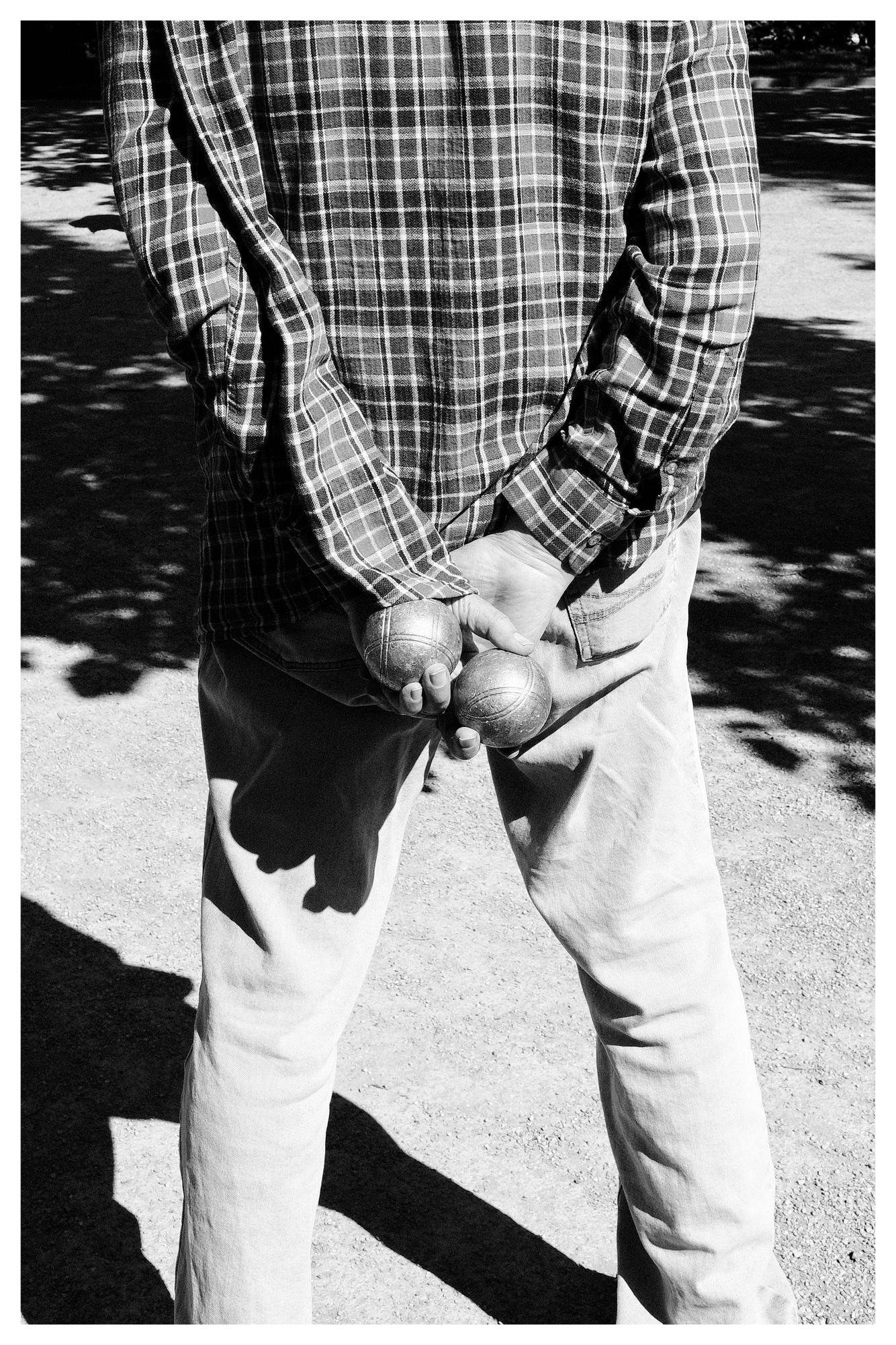 Black-and-white photo of a person in a plaid long-sleeve shirt and light pants standing on a gravel surface, holding two metal boule balls behind their back with both hands.