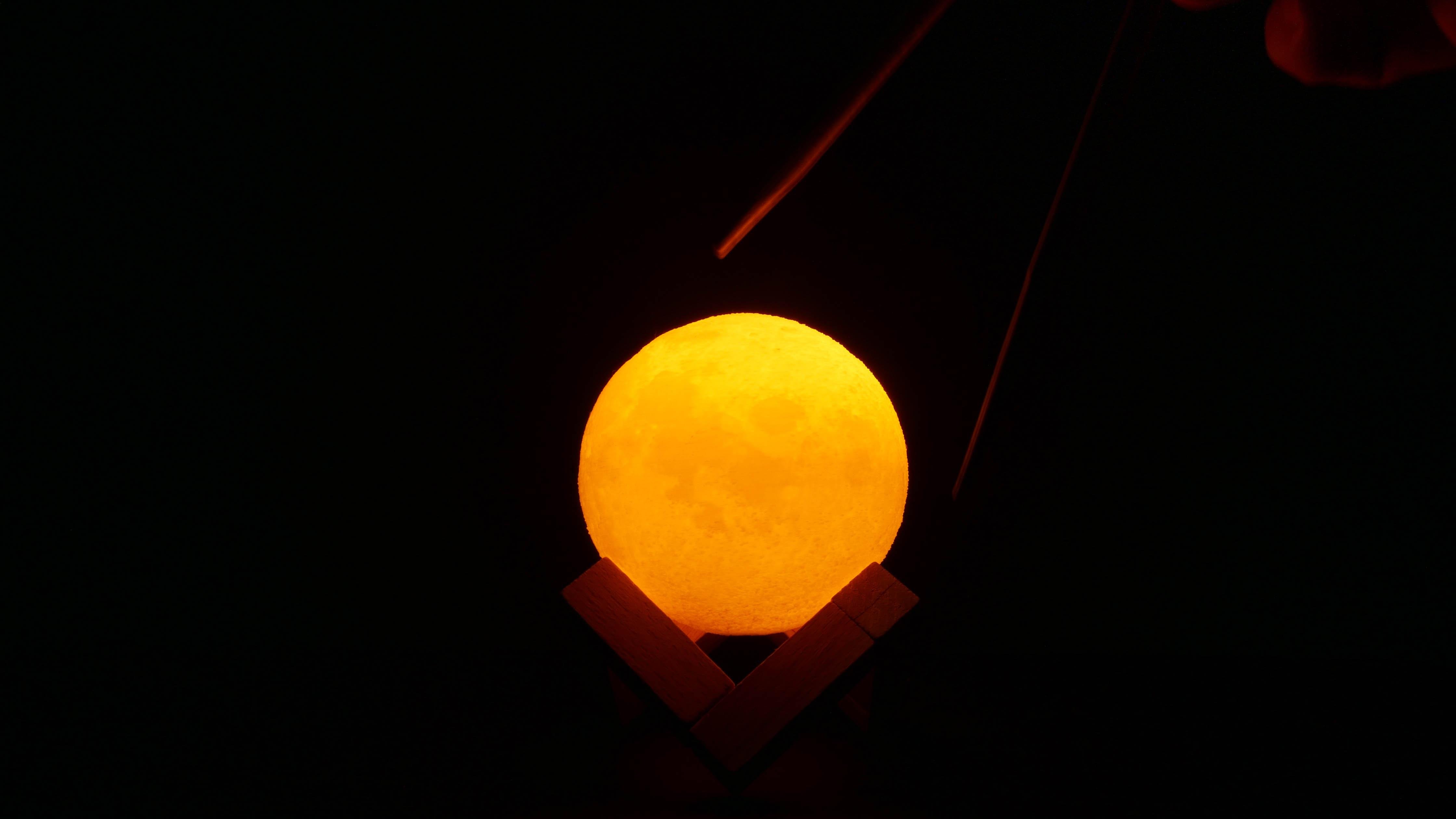 The image shows a glowing yellow sphere that resembles a moon lamp, positioned on a stand against a dark background. There appears to be a hand holding a pair of chopsticks near the lamp.