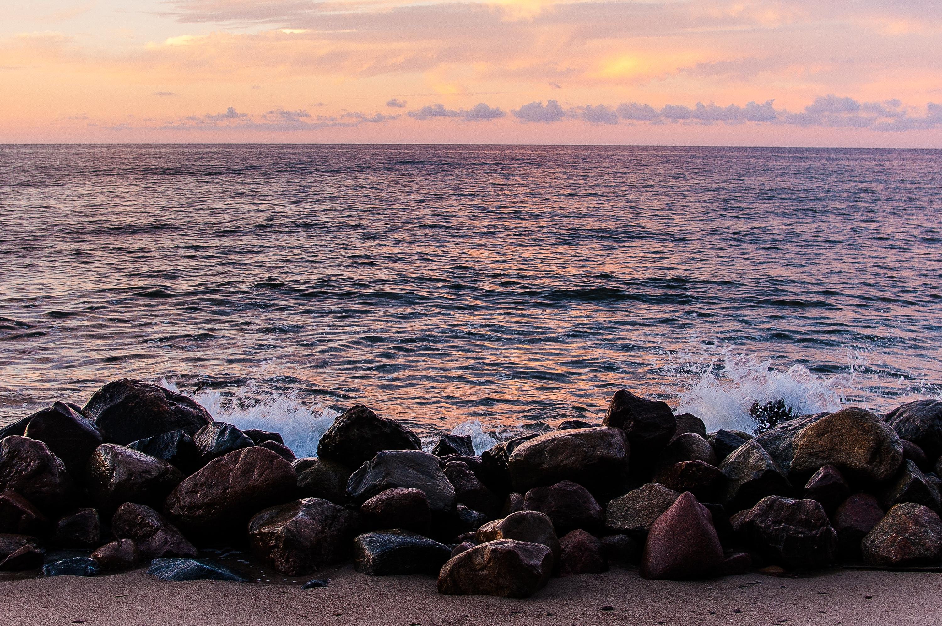 Coastal scene at sunset, sea with gentle waves. In the foreground, there are large rocks or boulders along the shore. The sky is a mix of soft pink and orange hues.