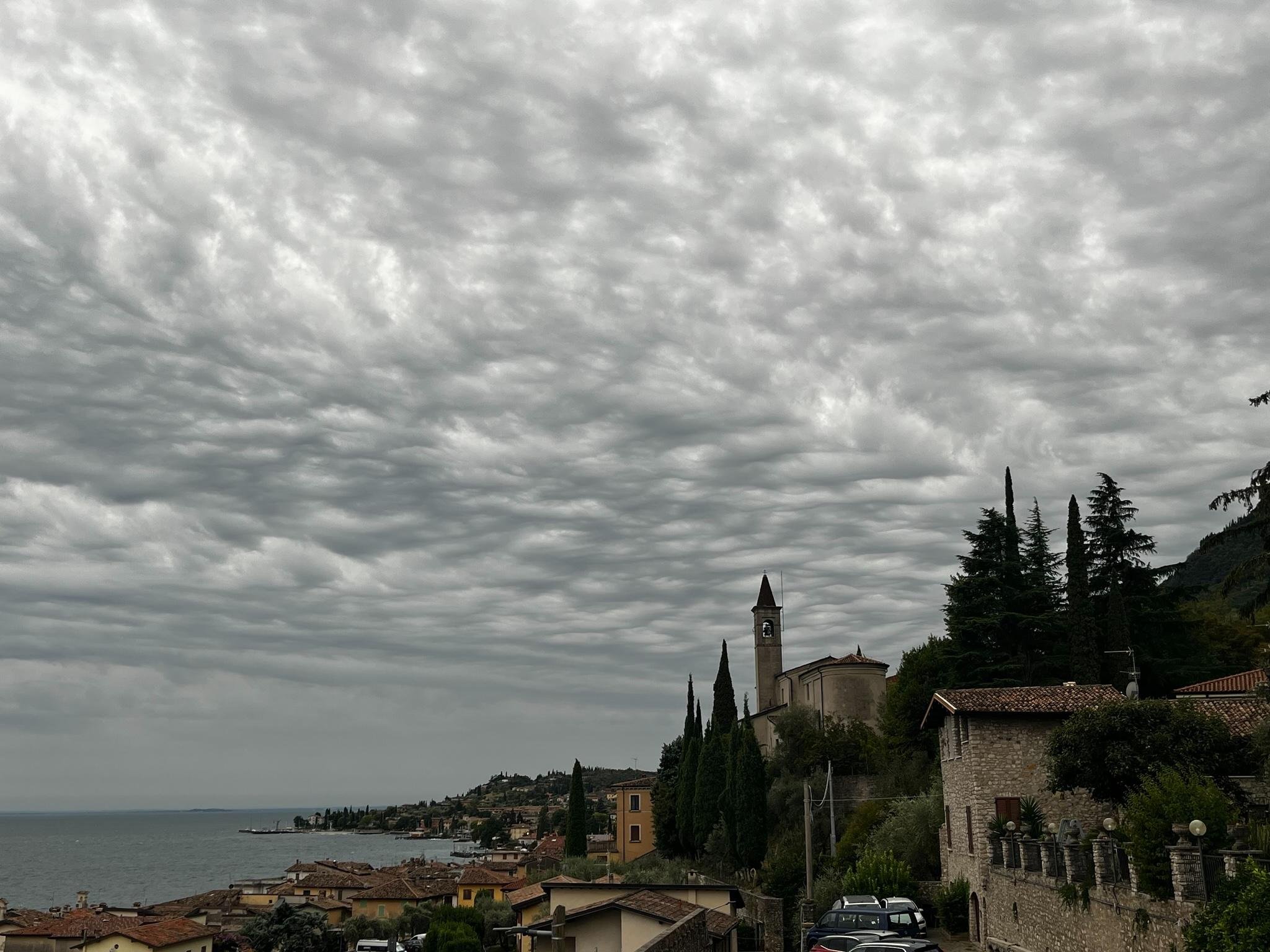 Coastal hillside town with terracotta rooftops beside a large lake, a stone church with a bell tower and cypress trees on the right, and a wide sky filled with thick gray clouds, similar to mammatus clouds, overhead.