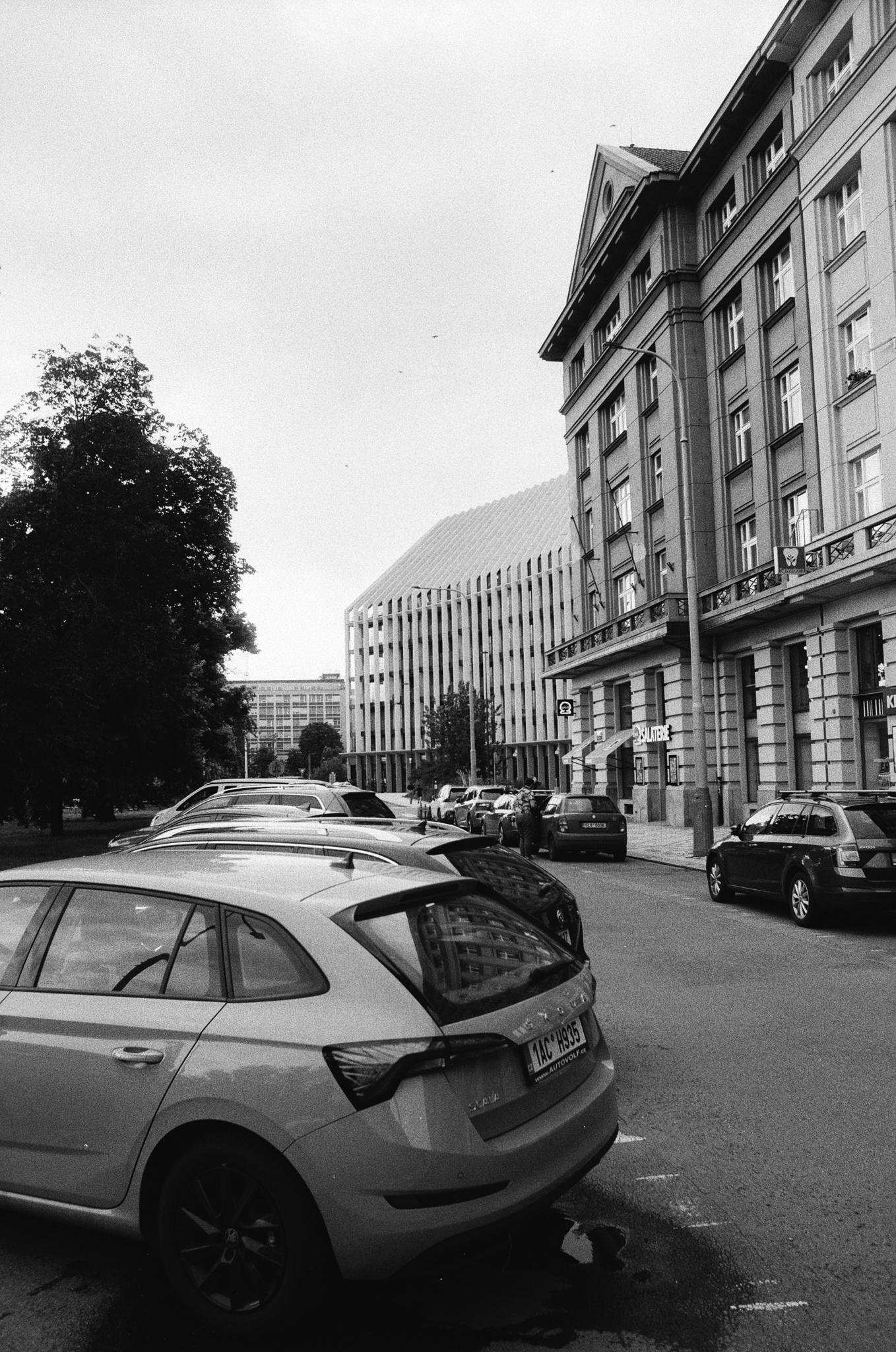 Black-and-white street scene with parked cars lining the curb in the foreground, a multi-story building with balconies and storefronts on the right, a large tree on the left, and a modern office block in the distance under an overcast sky.