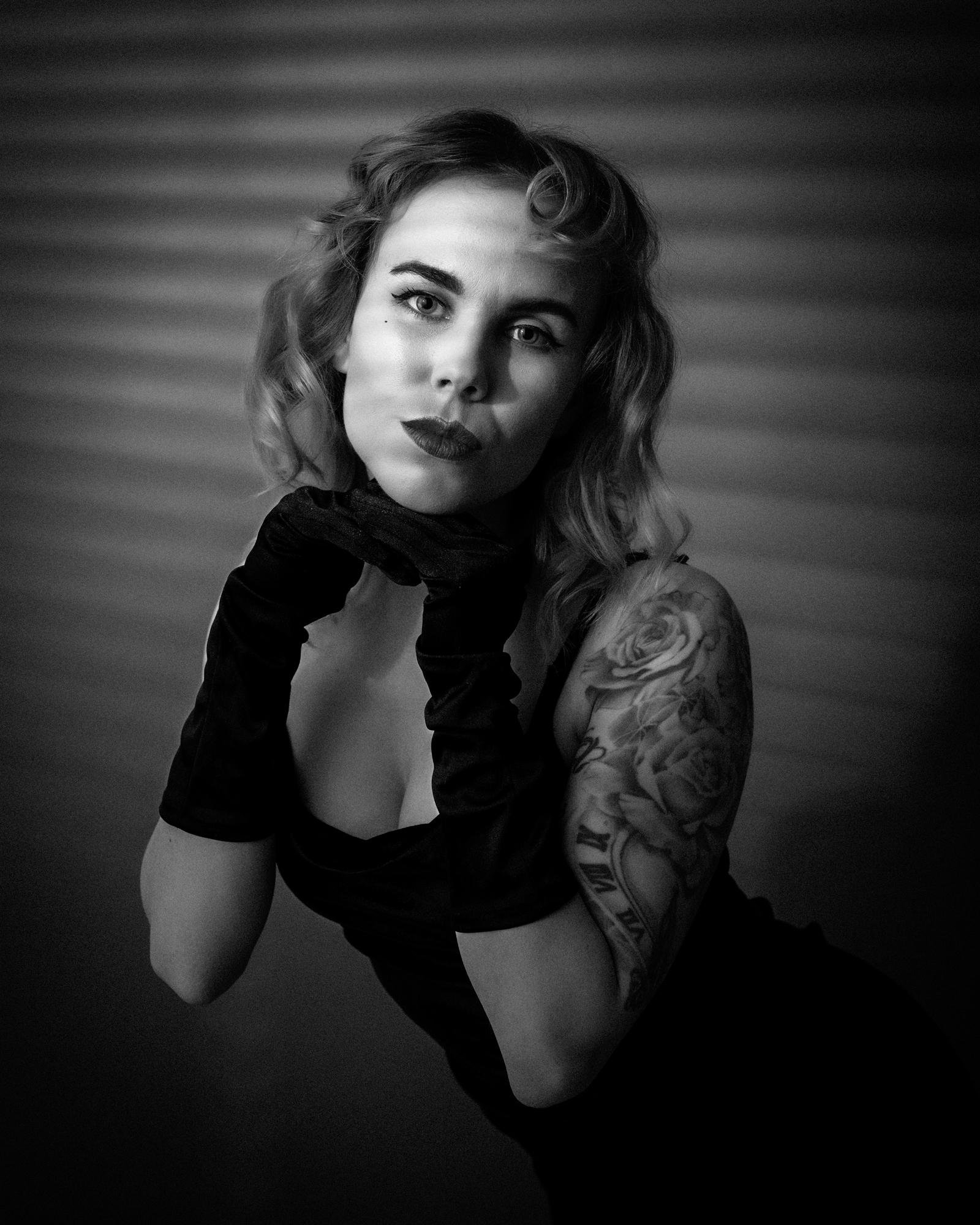 The image is a black and white portrait of a person with wavy hair, dark gloves, and a sleeveless outfit. They are posing with their hands under their chin. The person has a tattoo on their arm featuring roses and Roman numerals. The lighting and style give it a vintage or dramatic feel.