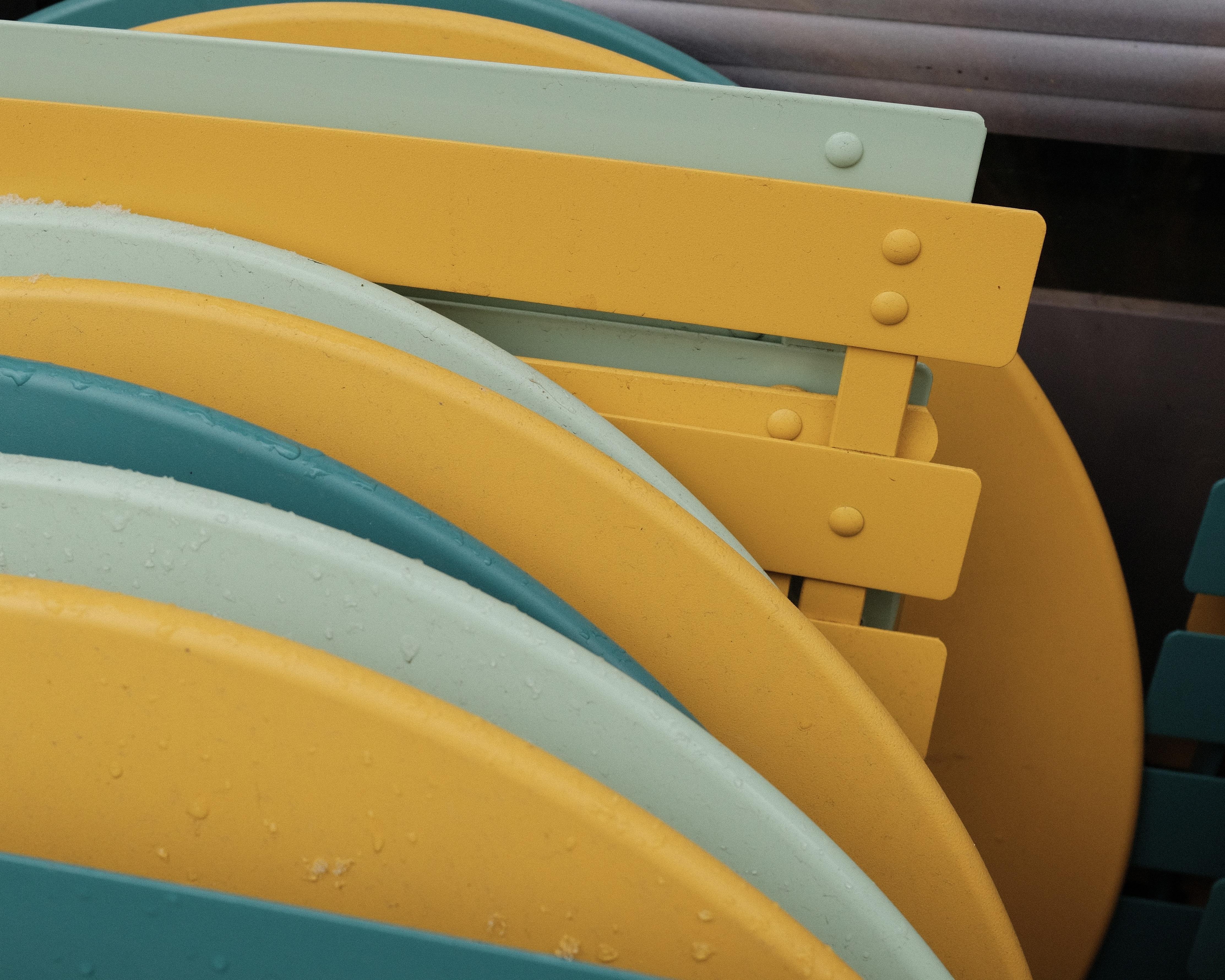 A closeup of stacked outdoor chairs and tables, as often seen in front of closed Cafés. The furniture items have bright yellow, petrol and light green colors. Since they are stacked, there are many layers of popping colors.