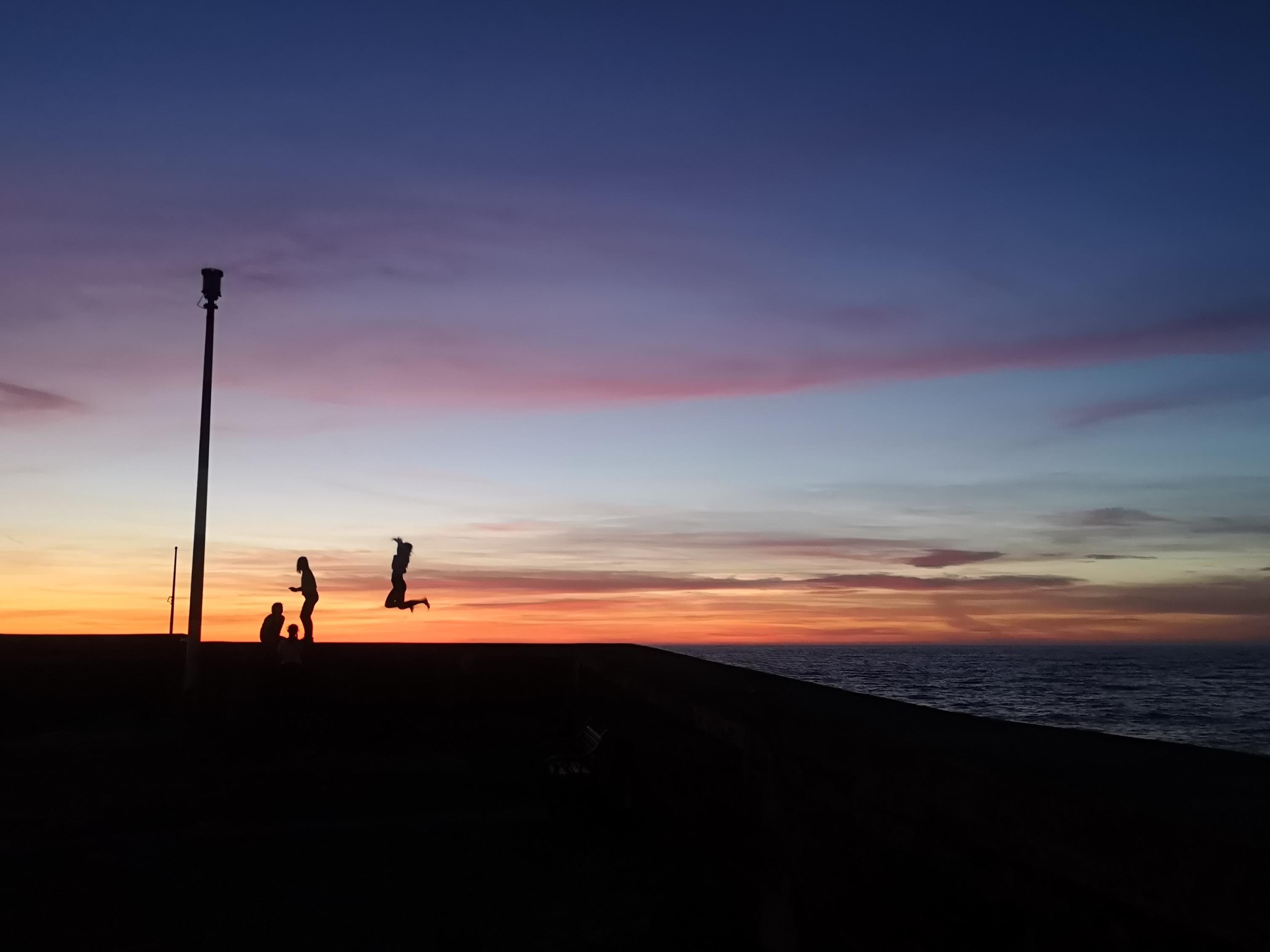 Silhouettes of a family on a harbour edge at sunset, with one person jumping, a tall pole on the left, and the ocean along the right under a purple-orange sky.