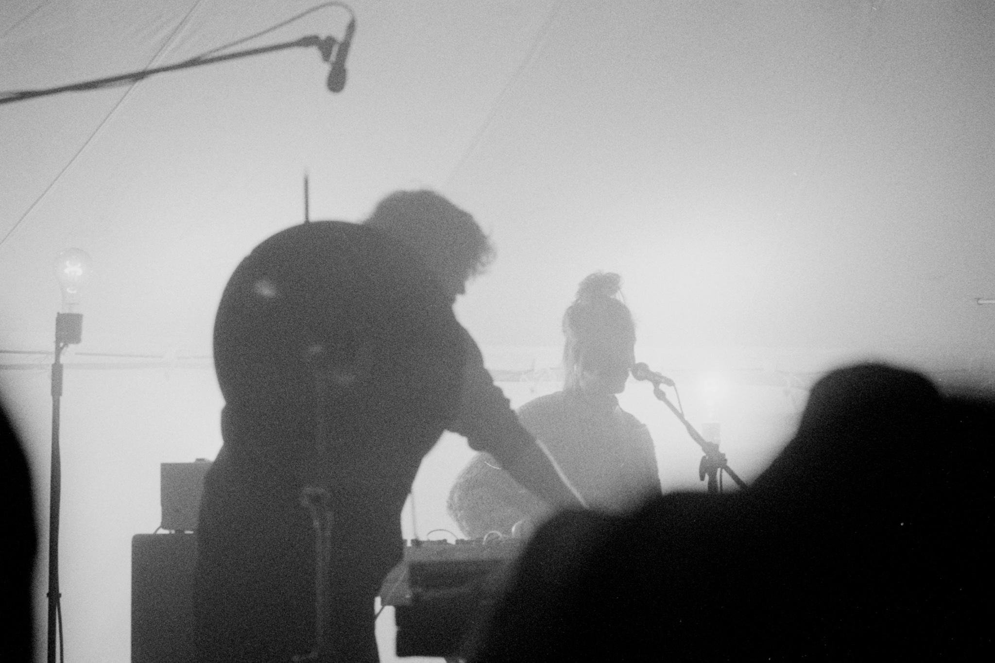 Silhouetted musicians performing on a dimly lit stage, with one person leaning over equipment in the foreground and another at a microphone; overhead boom mic, cables, and stage lights visible through haze.