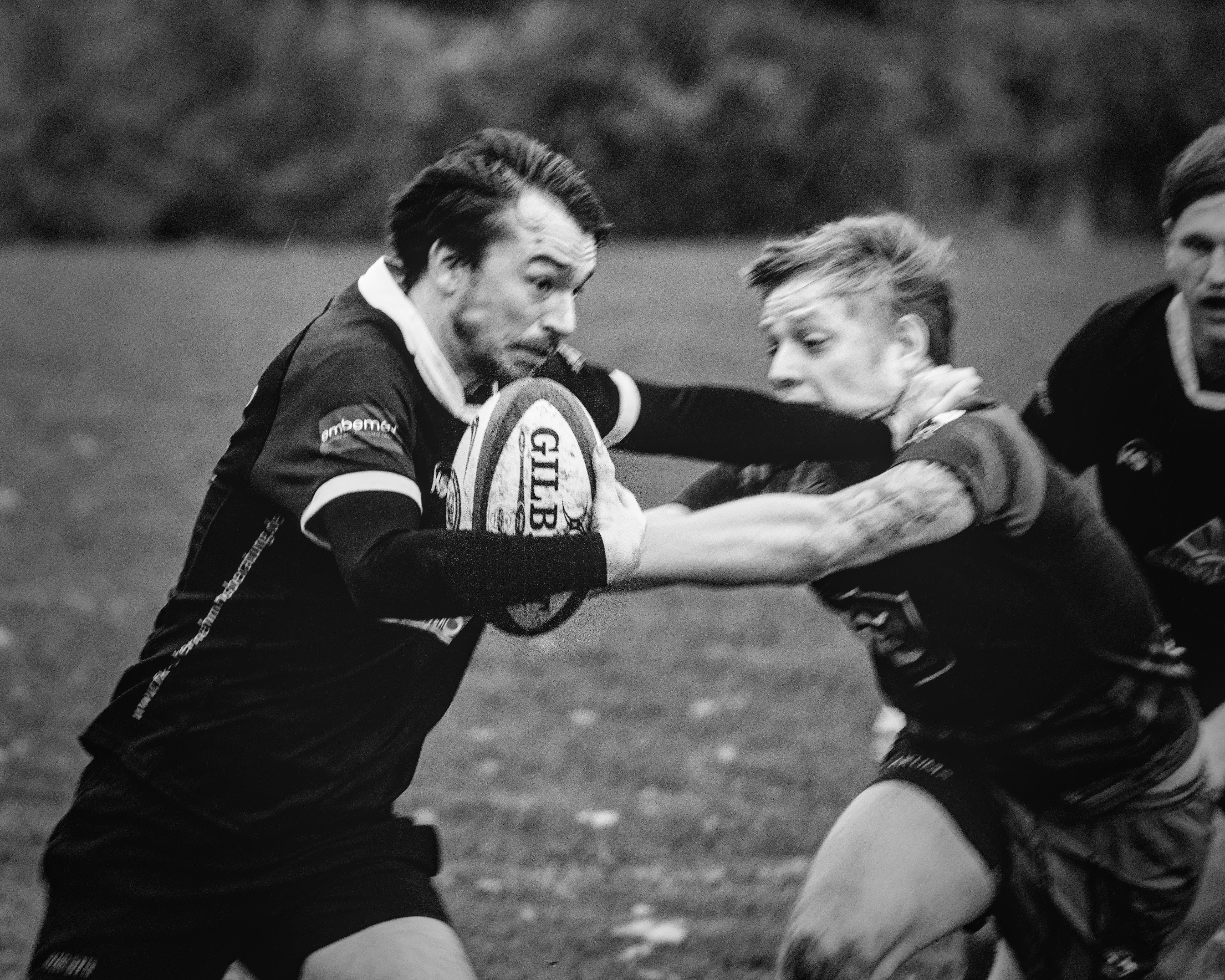 Two rugby players in action on a grass field. The player on the left, holding a ball marked "Gilbert," extends his arm to fend off the player on the right, who is reaching out. Both are wearing dark jerseys. The background is blurred, highlighting the players' movement and focus.
###
Zwei Rugbyspieler in Aktion auf einem Rasenfeld. Der Spieler auf der linken Seite, der einen Ball mit der Aufschrift "Gilbert" hält, erweitert seinen Arm, um den Spieler auf der rechten Seite abzuwehren, der die Hand ausstreckt. Beide tragen dunkle Trikots. Der Hintergrund ist verschwommen und unterstreicht die Bewegung und den Fokus der Spieler.