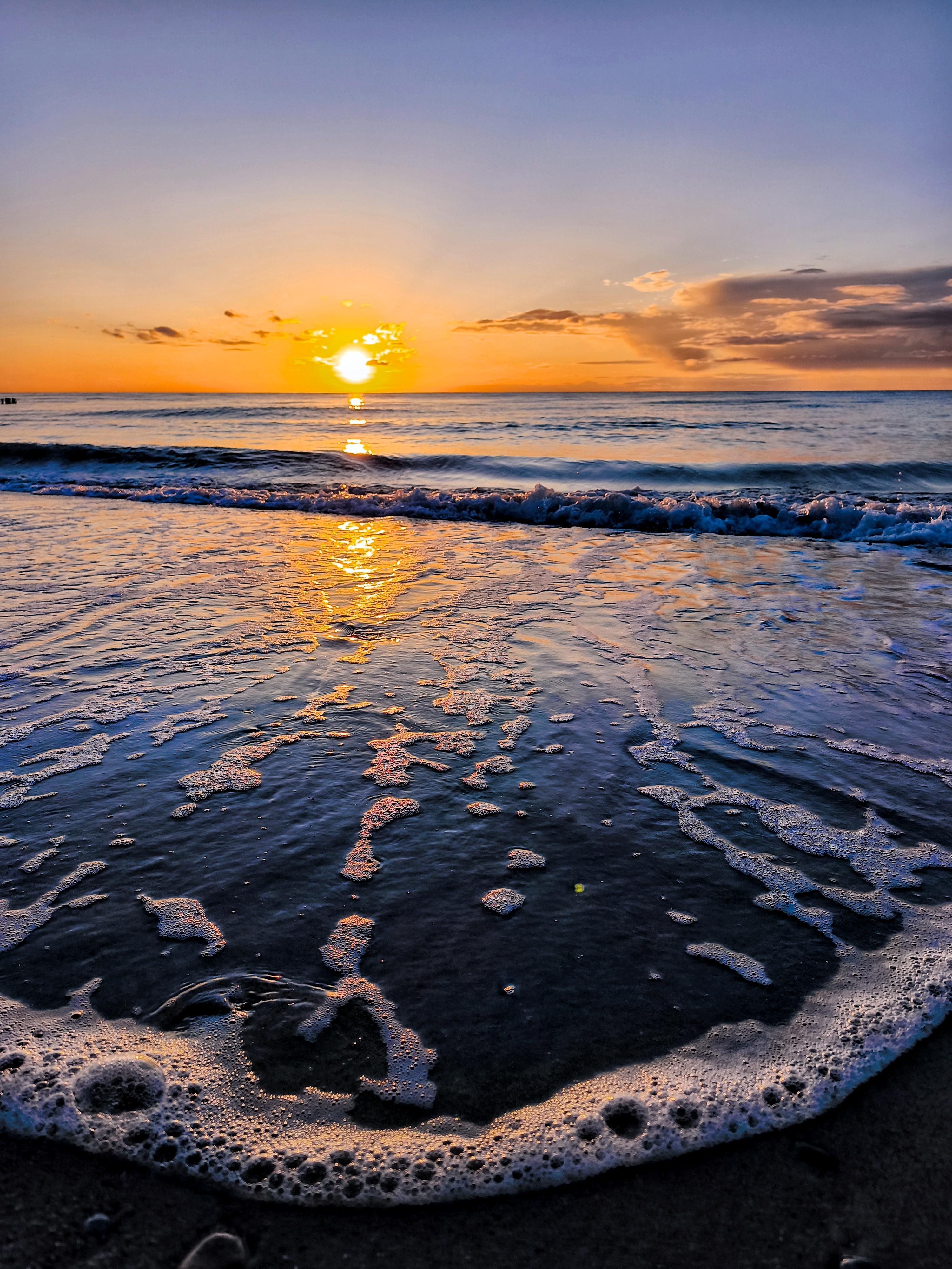 Low-angle view of a beach at sunset, with foamy surf washing over dark wet sand in the foreground and the sun near the horizon reflecting a golden path across calm ocean water under scattered clouds.