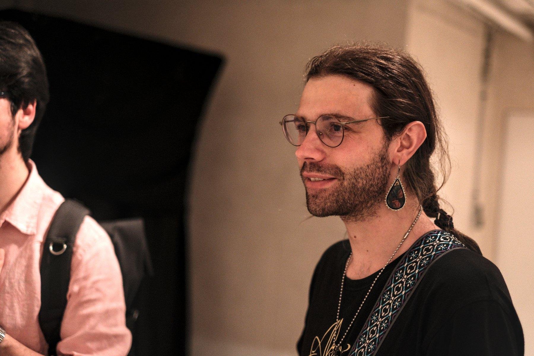 Photo horizontale. Un homme brun aux cheveux longs attaché derrière lui, lunettes et une boucle d’oreille en forme de goutte, souriant en regardant devant lui. À gauche, coupé par le cadre, un autre homme brun en chemise rose