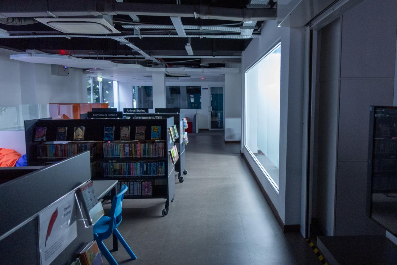 Dimly lit library interior with rows of bookshelves containing various books, labeled sections "Humorous Stories" and "Animal Stories." A blue chair is positioned near the shelves. The right side features a large window, and the ceiling has visible ducts and lights. The hallway extends into the distance, illuminated by ambient light.