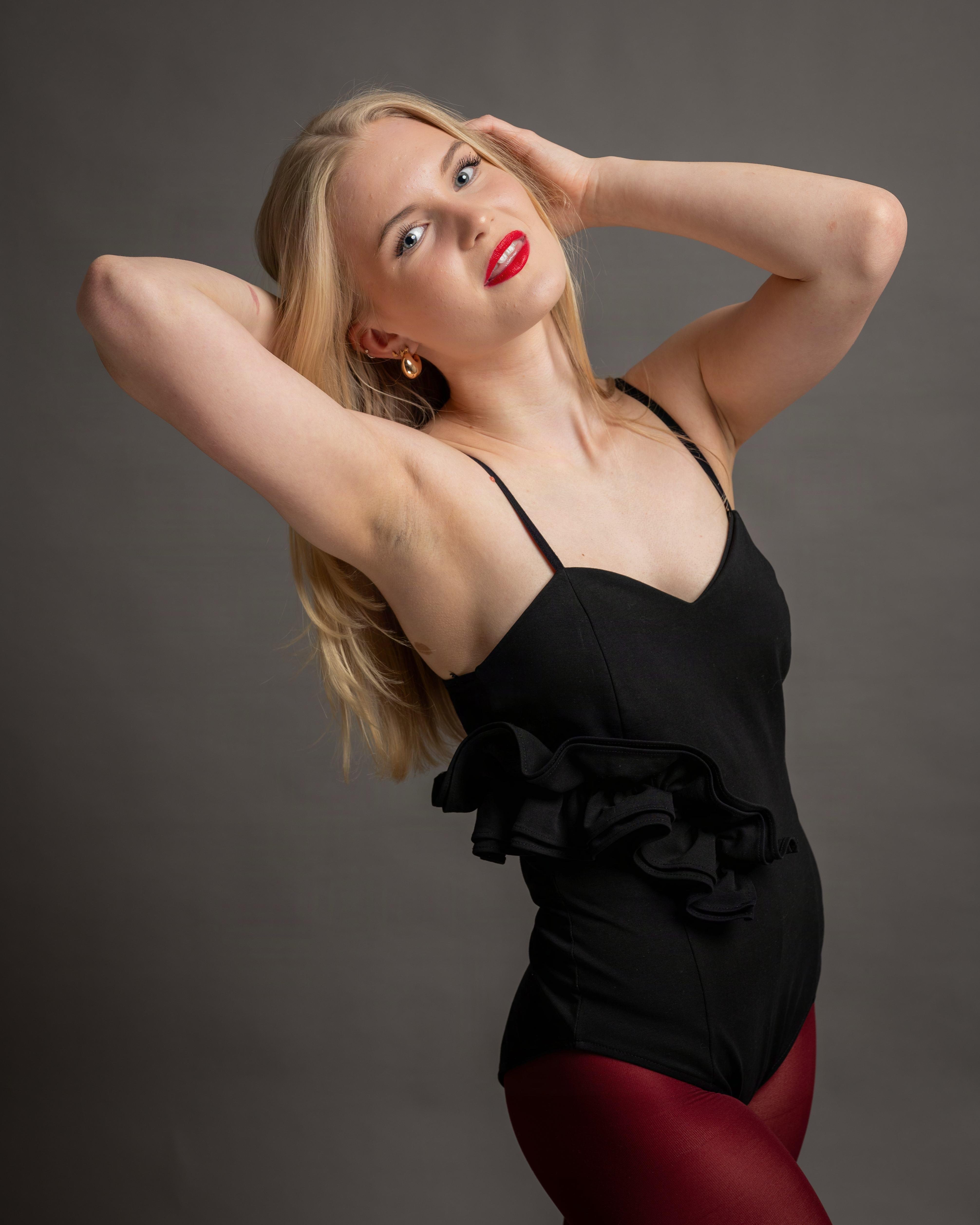 Woman with long blonde hair wearing a black outfit with thin straps and ruffled detailing, paired with red leggings. Her arms are raised, with hands touching her head. She has red lipstick and is against a plain gray background.