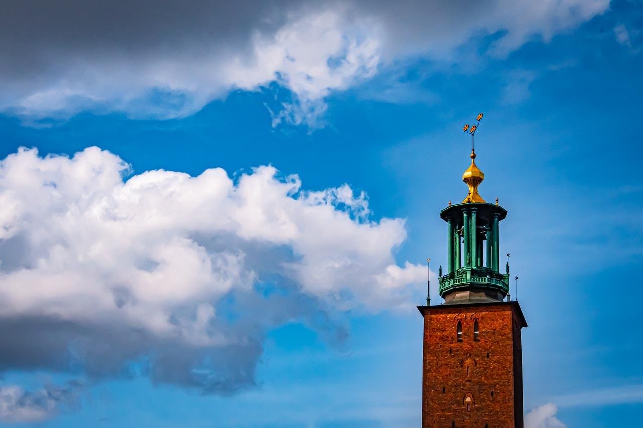 Brick tower with a green and gold ornate spire, set against a vibrant blue sky with scattered white clouds. The spire features a weather vane at the top.