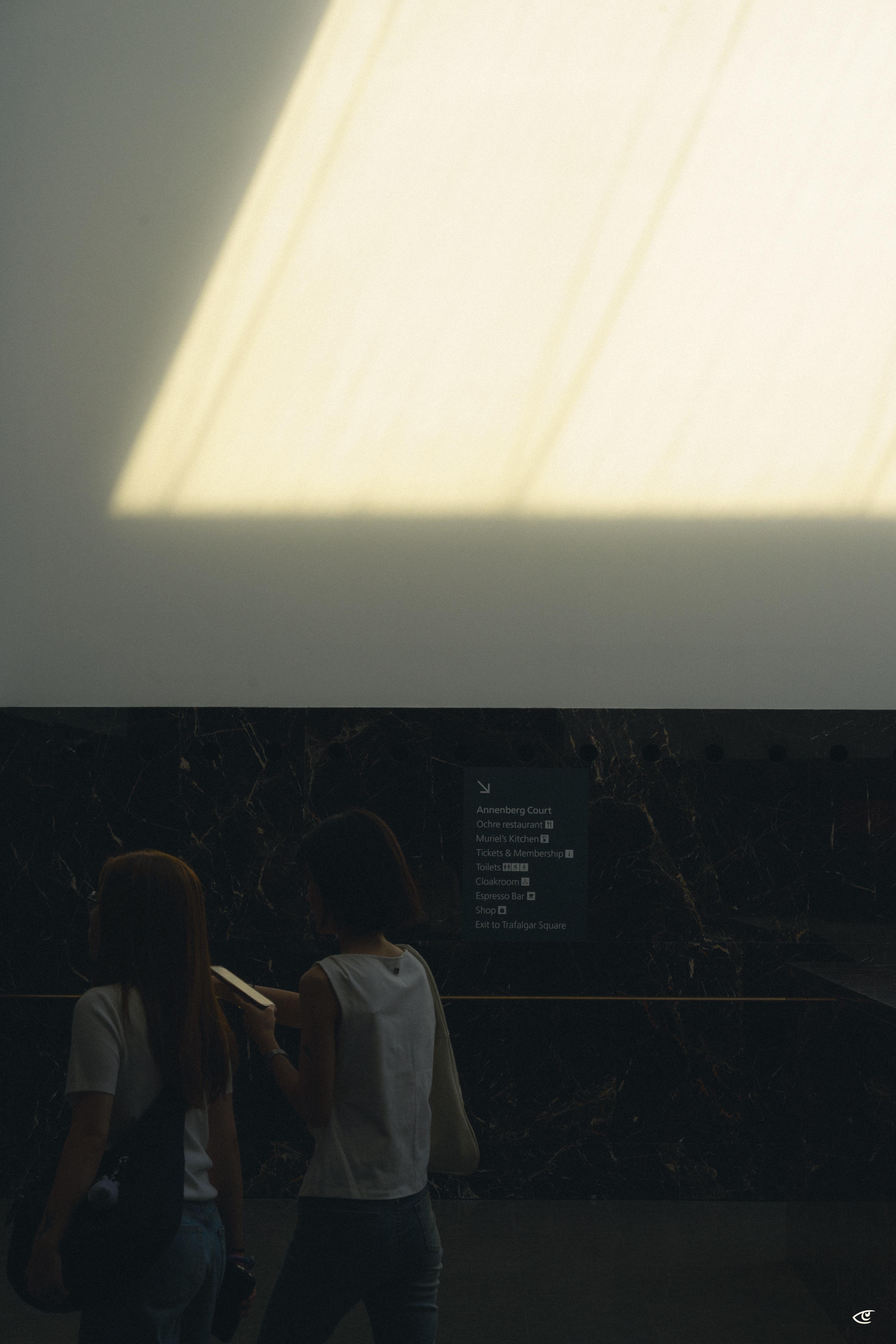 Two people seen from behind in a dim interior, standing near a dark marble wall with a directory sign, while bright sunlight from a skylight casts a large rectangular patch of light and shadow across the upper wall.