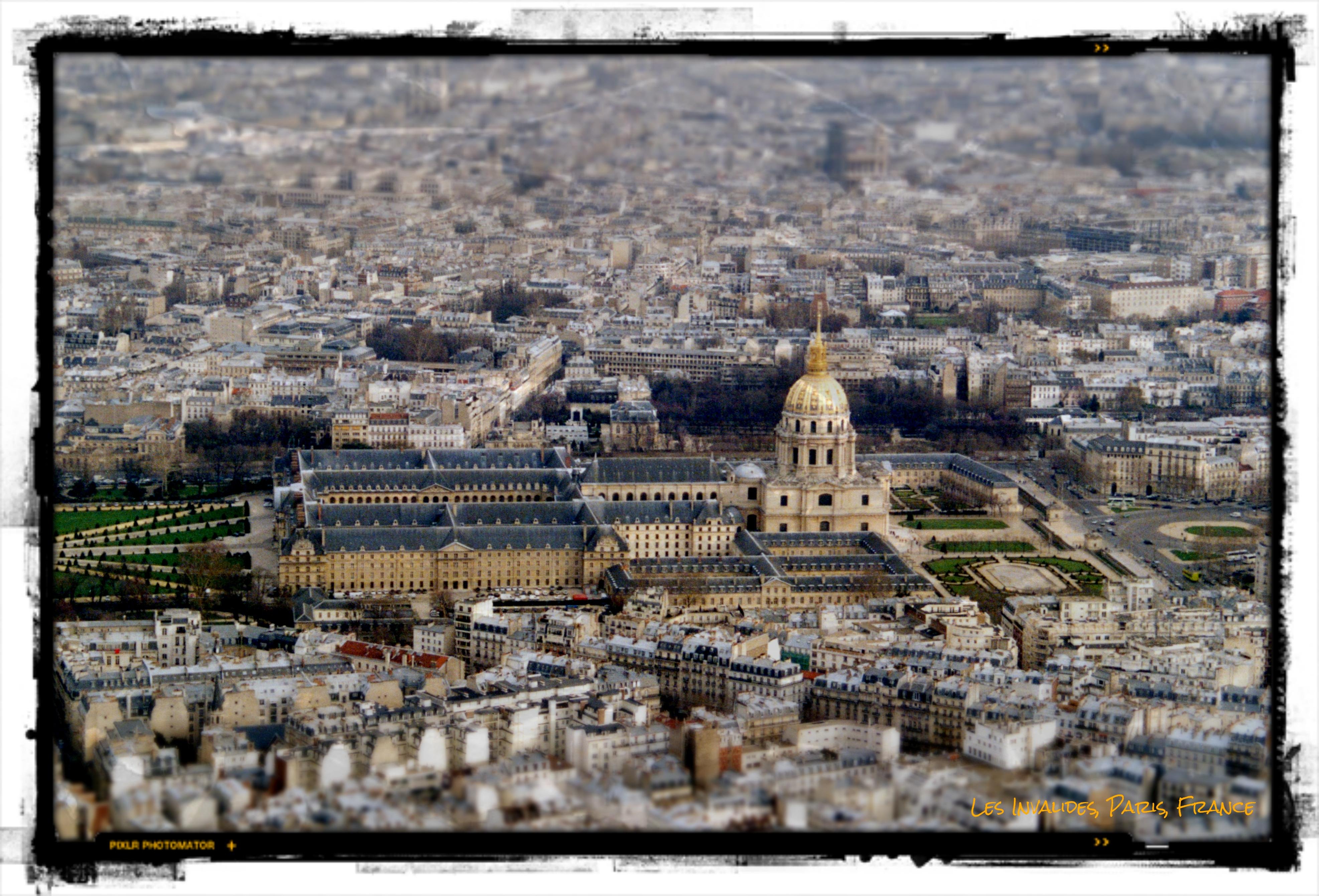 Digitized and edited scan of a 35mm film photograph taken in February 2004 from the observatory of the Eiffel Tower, overlooking the historic complex of Les Invalides in Paris. The image has a slightly soft, grainy quality due to the limitations of the original scanner, with subtle tilt-shift–like blur toward the edges. At the center stands the striking gold dome of the Dôme des Invalides, gleaming above the symmetrical stone buildings and courtyards of the Hôtel des Invalides. Surrounding it stretches a dense expanse of Parisian rooftops, pale grey and cream, forming an intricate urban tapestry fading into the atmospheric haze in the background. A black, distressed border frames the image, with small yellow text in the lower right reading “Les Invalides, Paris, France,” and faint branding from Pixlr Photomator appearing on the lower left.