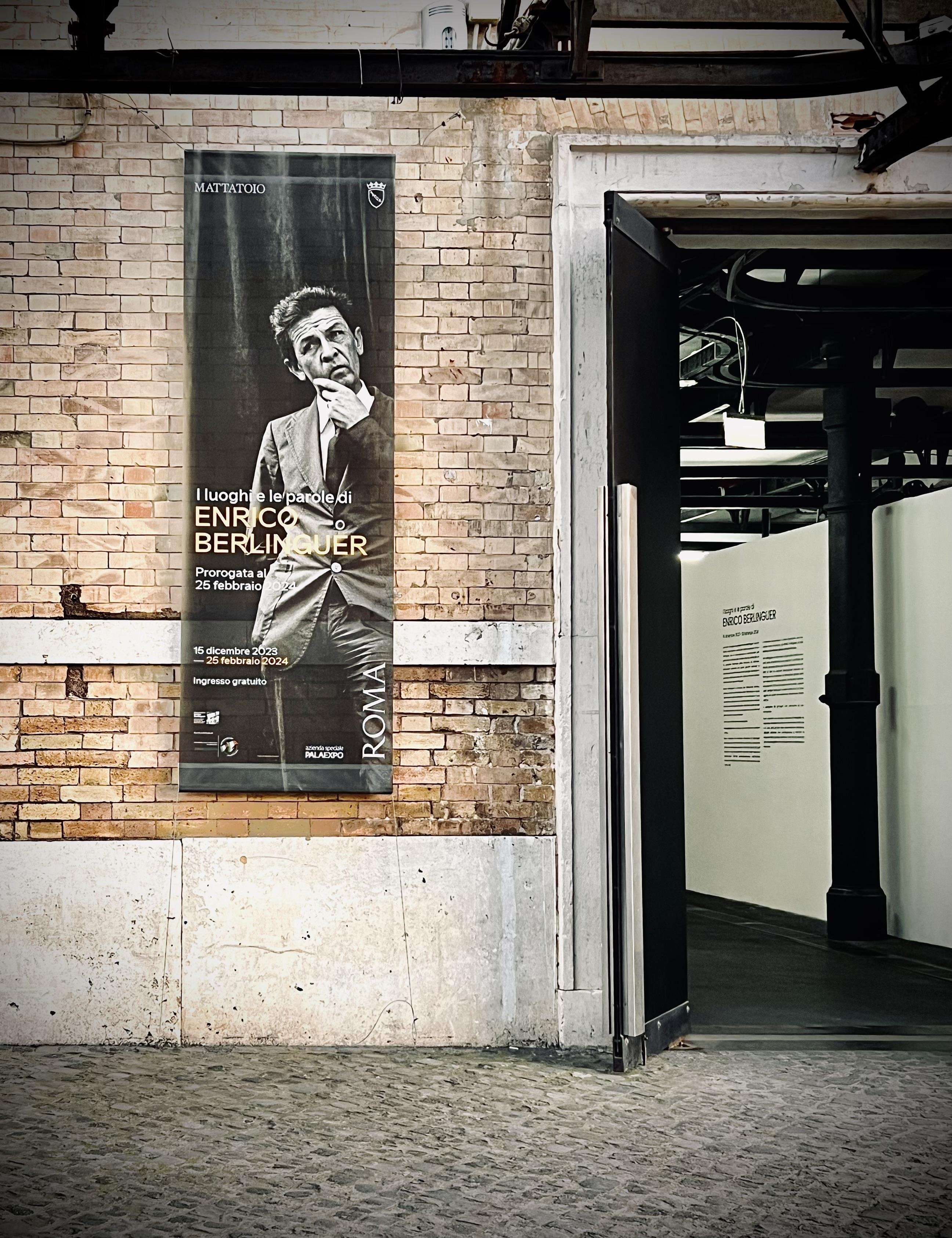 Poster su un muro di mattoni. Il poster presenta una fotografia in bianco e nero di un uomo e include un testo che recita "I luoghi e le parole di Enrico Berlinguer," indicando una mostra o un evento sui luoghi e le parole di Enrico Berlinguer. L'evento si svolge dal 15 dicembre 2023 al 25 febbraio 2024 ed è gratuito. La location è a Roma, in un luogo chiamato "MATTATOIO". Accanto al poster c'è una porta aperta che conduce a uno spazio espositivo nel muro.