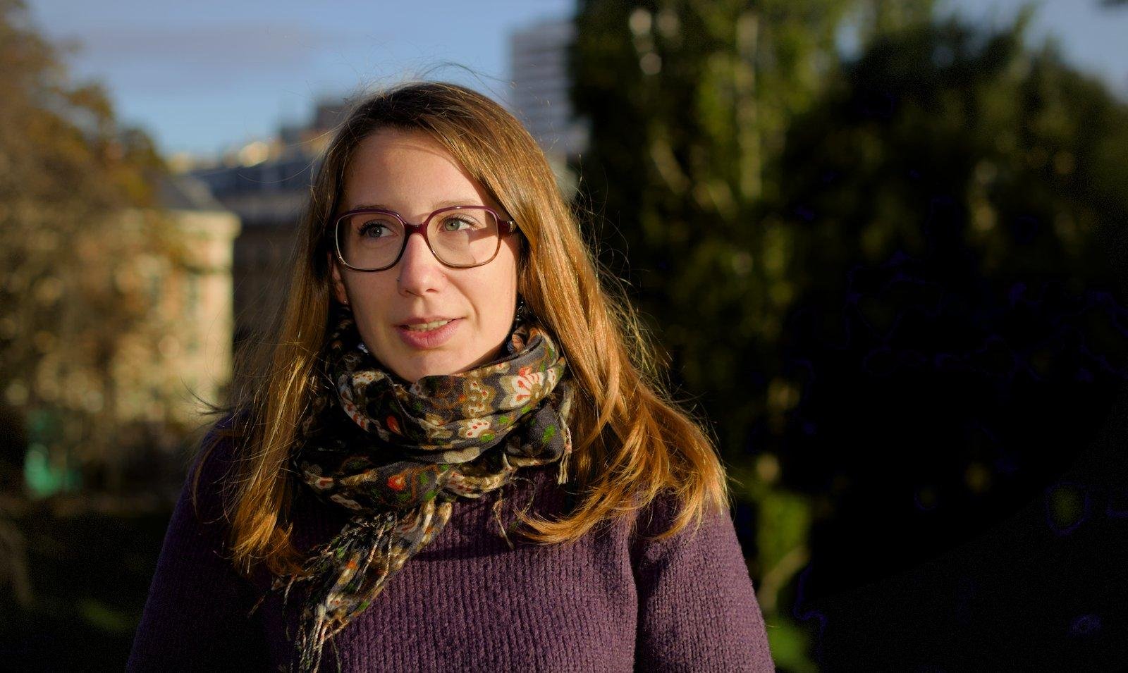 Photo format paysage montrant Une fille châtain de face mais avec le regard vers la gauche de la photo, une lumière chaude de type levé de soleil l’éclaire par la droite de l’image. Le fond est plein d’arbres et on distingue un peu d’immeubles parisiens