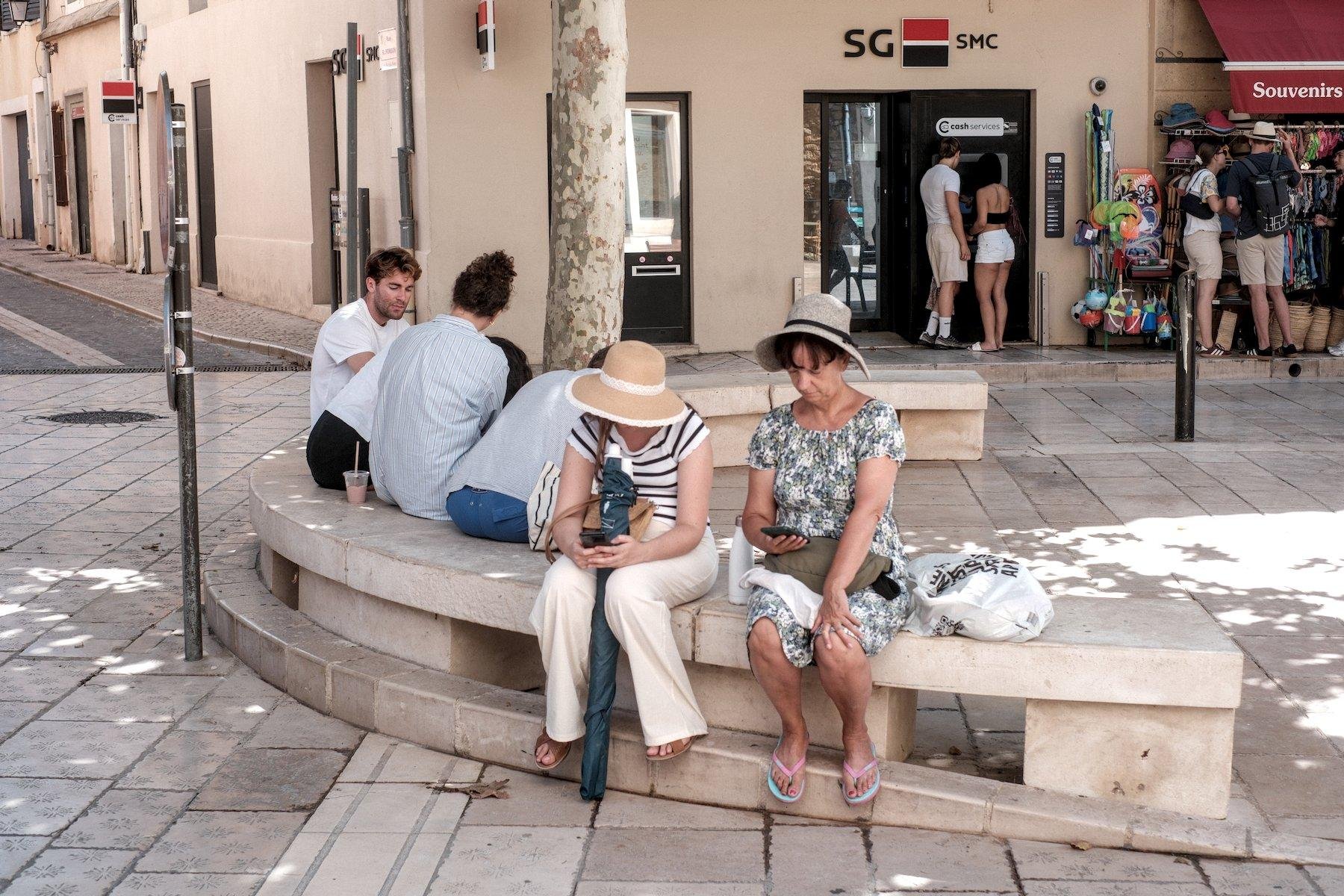 Horizontal photo. In the street, a public bench made of concrete paint in white, in a half circle shape, with 2 groups of people on it, in inverted sides, 4 young (ish) and 2 older ladies with hats