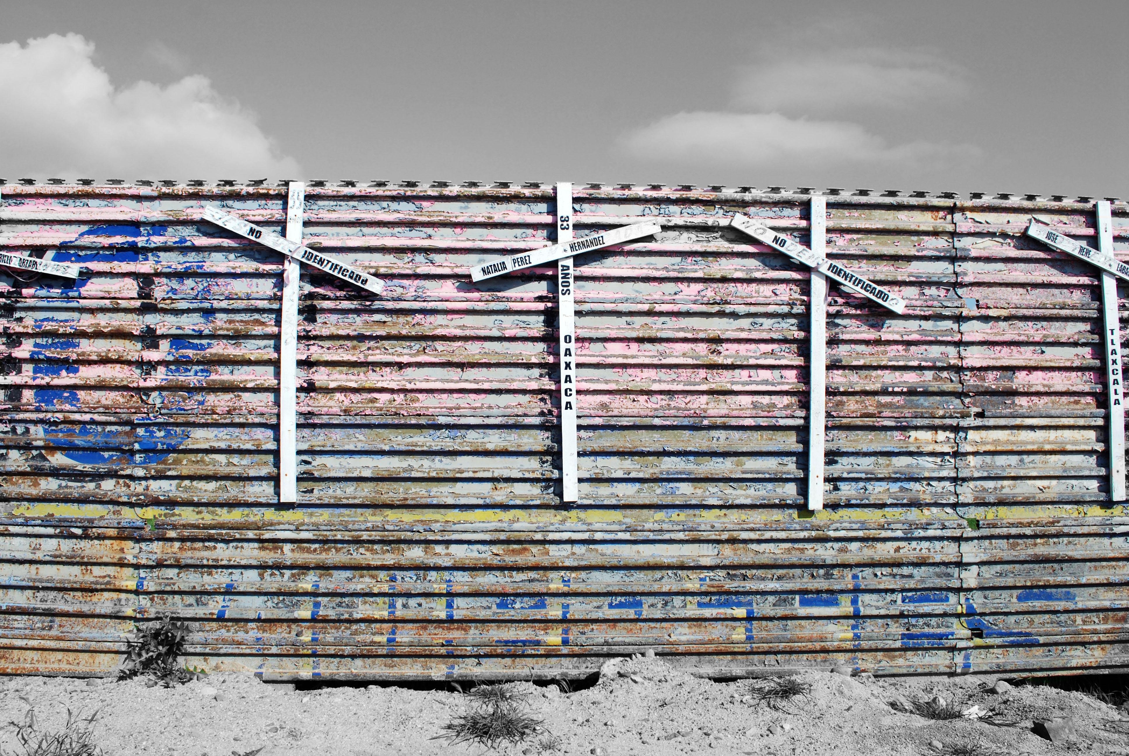 The image shows a section of a painted, corrugated metal fence or wall with crosses attached to it. The wall has various shades of paint, and the crosses have inscriptions on them. The ground in front of the fence is dry and sandy, indicating a desert-like environment. The sky is partly cloudy.