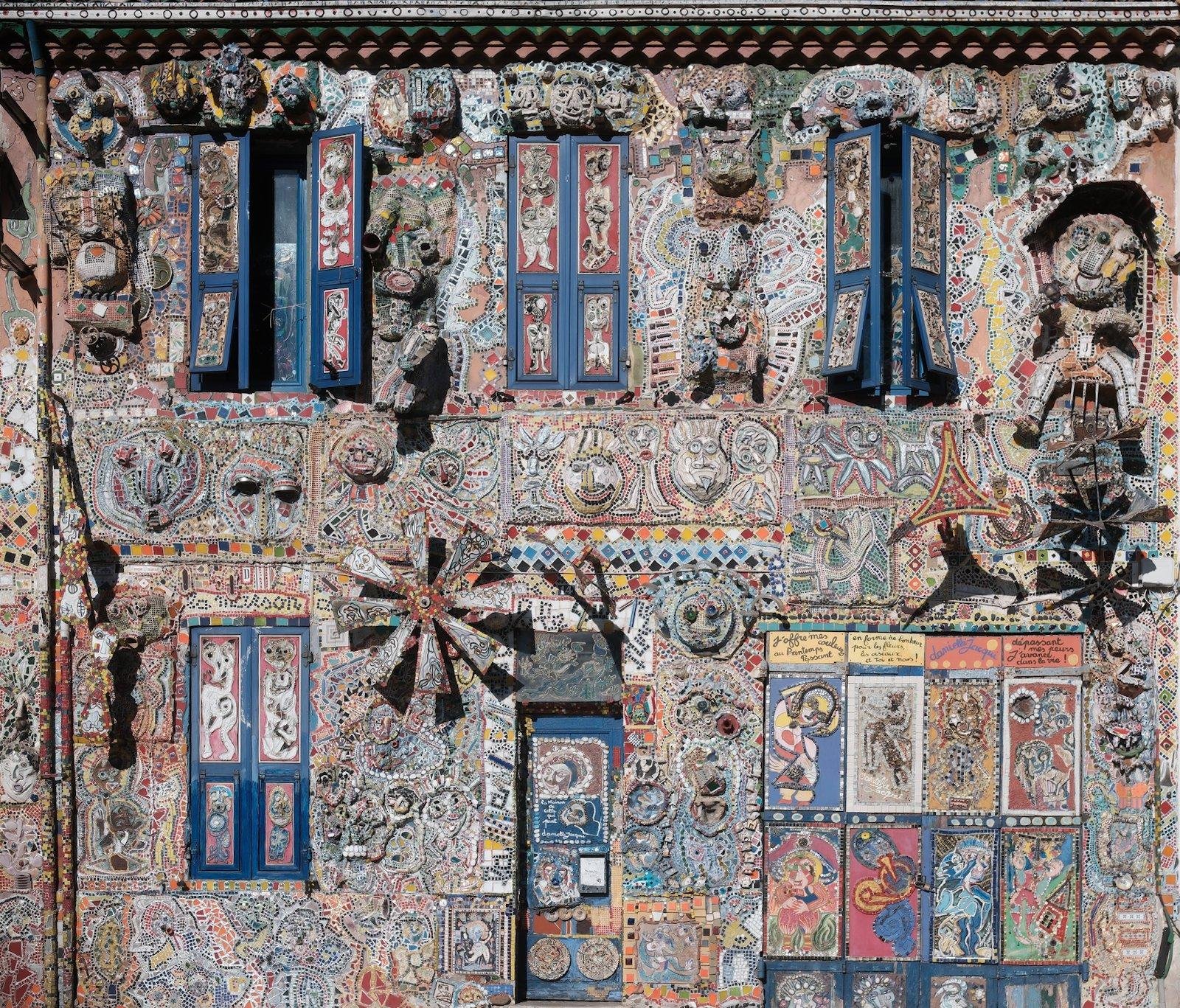 Photo de la façade d’une maison entièrement décorée de plusieurs milliers de céramiques qui forment des motifs tantôt abstraits, tantôt représentant quelque chose et bardée de décoration autres en plus de la céramique. ENTIÈREMENT couverte, même l’intérieur des volets.