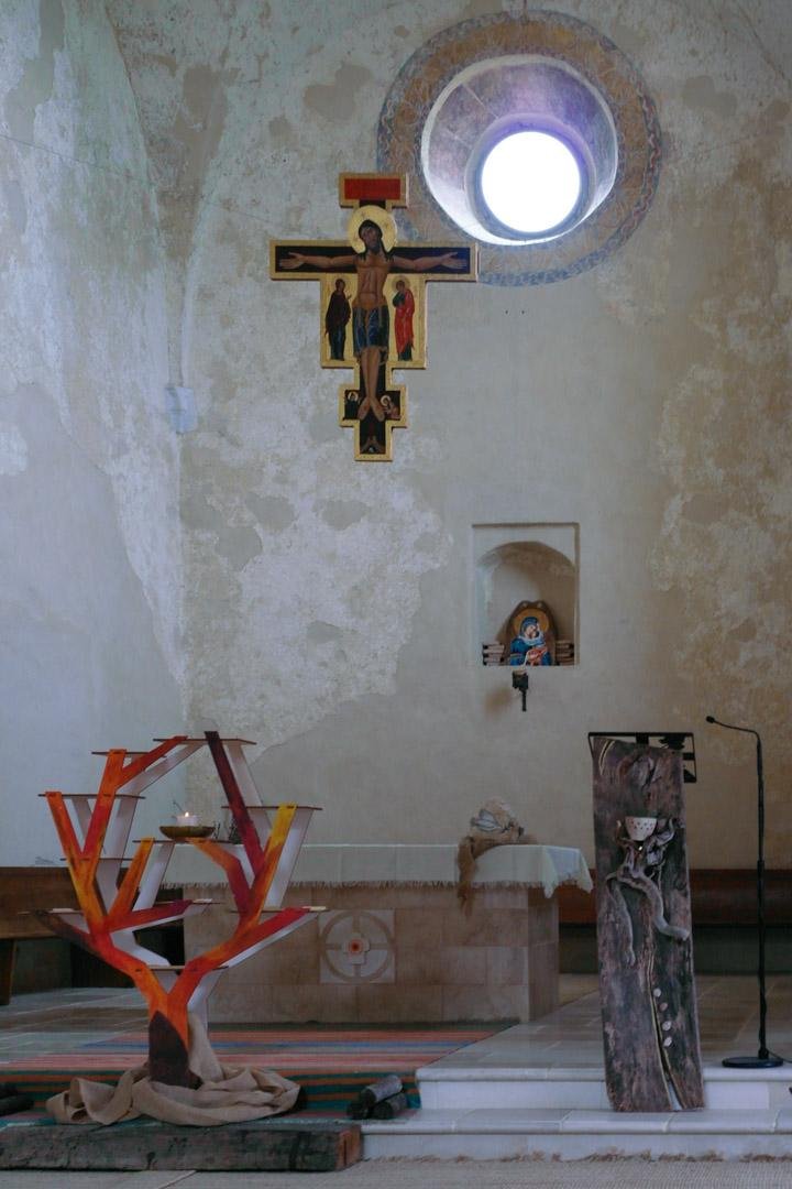 Interno di una chiesa spoglia, illuminata da una apertura circolare sullo sfondo. Un crocifisso pende dal soffitto, mentre in basso si vedono l'altare, il supporto per il leggio ricavato da un blocco di legno scolpito e un supporto per le candele simile a un albero spoglio.