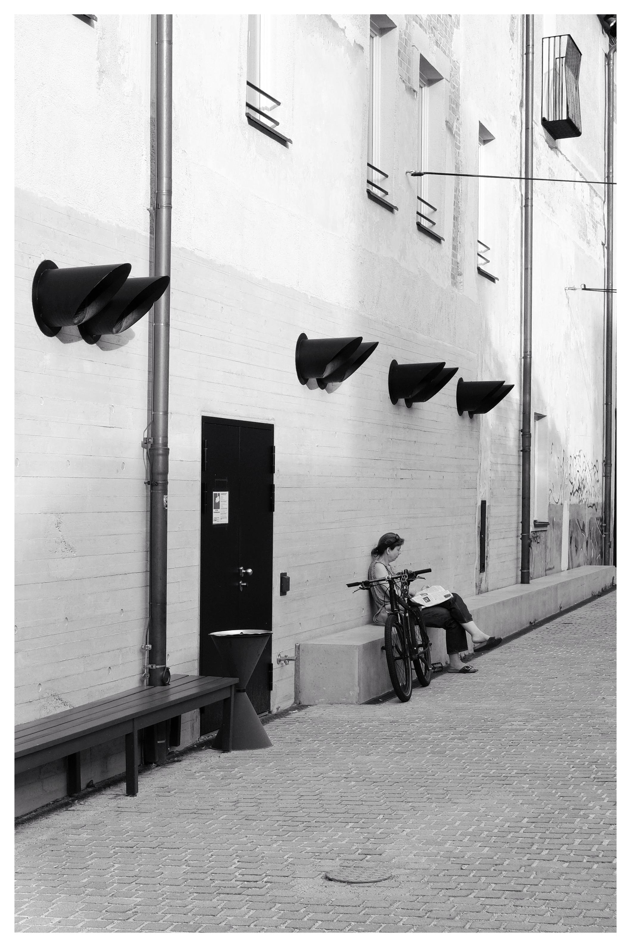 Black and white photo of an alleyway with a person sitting on a concrete bench reading a newspaper next to a bicycle. The wall features five large, angled black vent structures and several small windows with security bars. A dark door with a posted sign and metal drainpipe is visible on the left side. The ground is paved with small bricks.