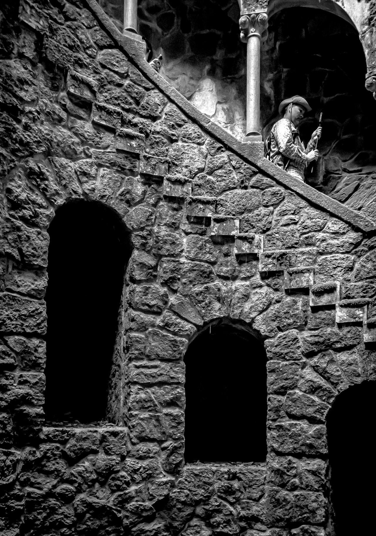 Stone spiral staircase with rough, textured walls featuring two arched openings. A person wearing a hat and holding a walking stick is visible near the top, partially obscured by a column. The scene is in black and white, highlighting the intricate patterns of the stonework.