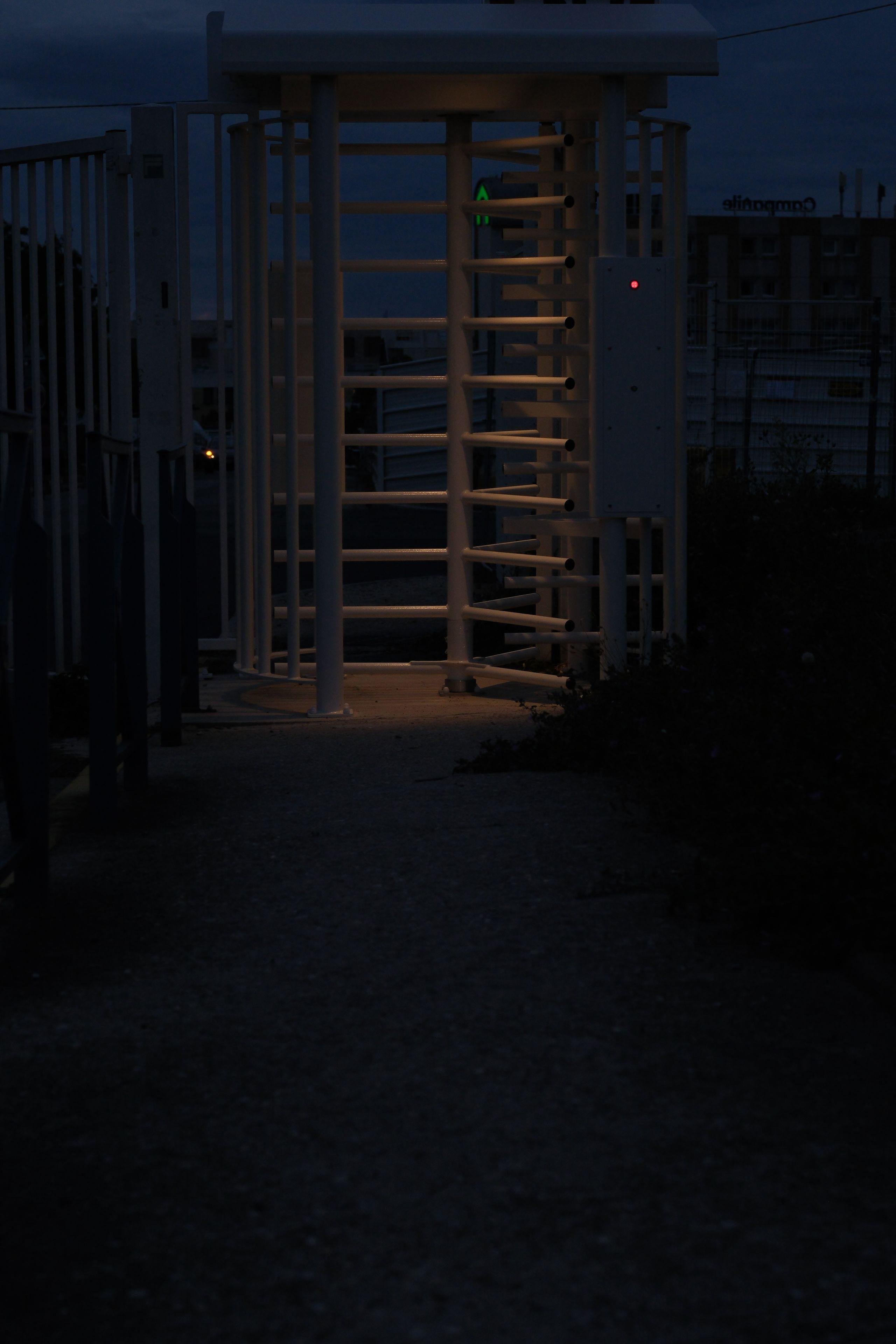 Photo format portrait de nuit, d’une porte blanche avec des barreaux horizontaux rotatifs. Elle est faiblement éclairée par une lumière orangée, sur fond de ciel bleu nuit