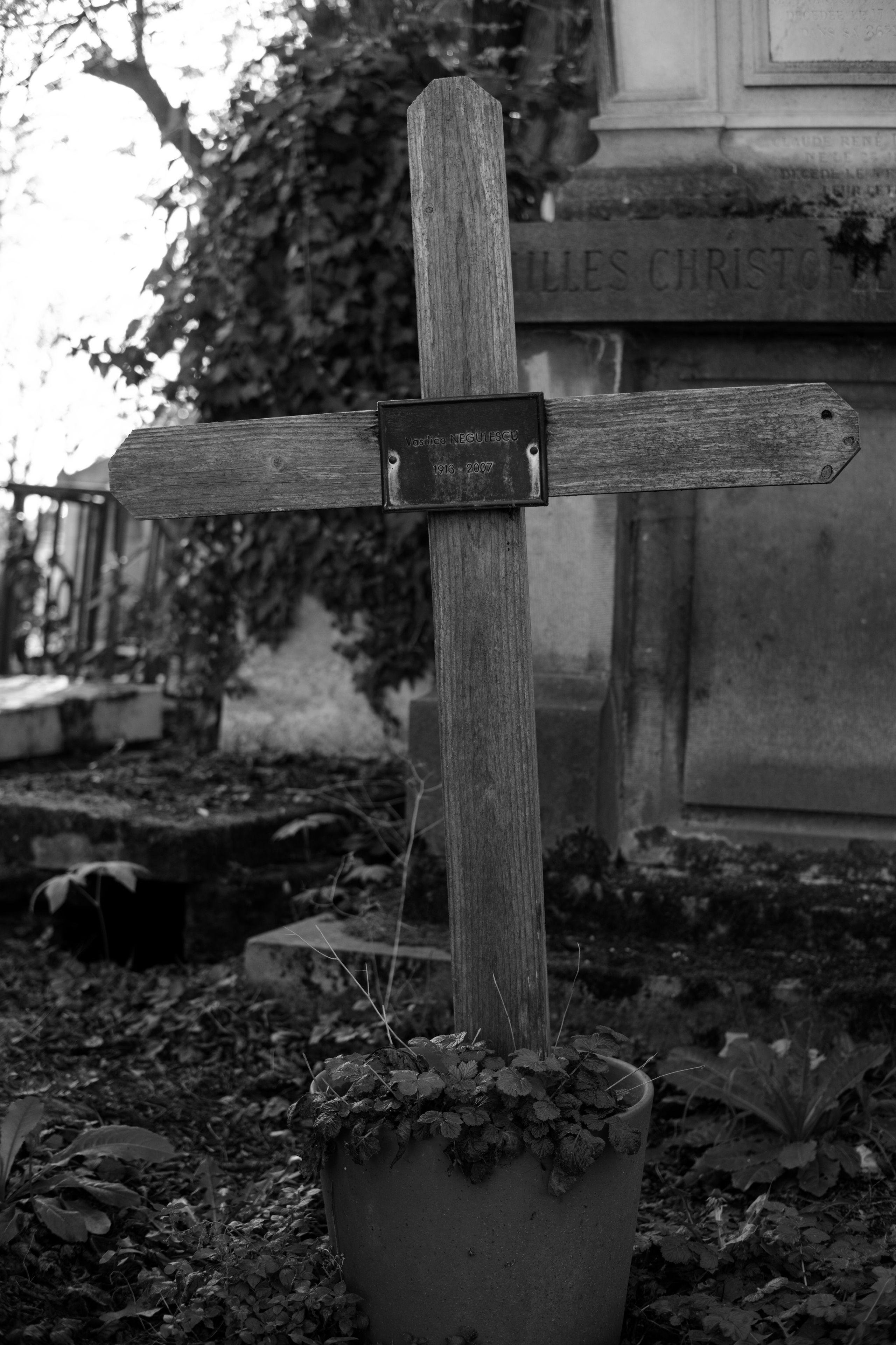 Photo verticale en noir et blanc. Une tombe faite d’un gros pot de fleur dans lequel est enfichée une croix en bois avec une petite plaque au centre de la croix