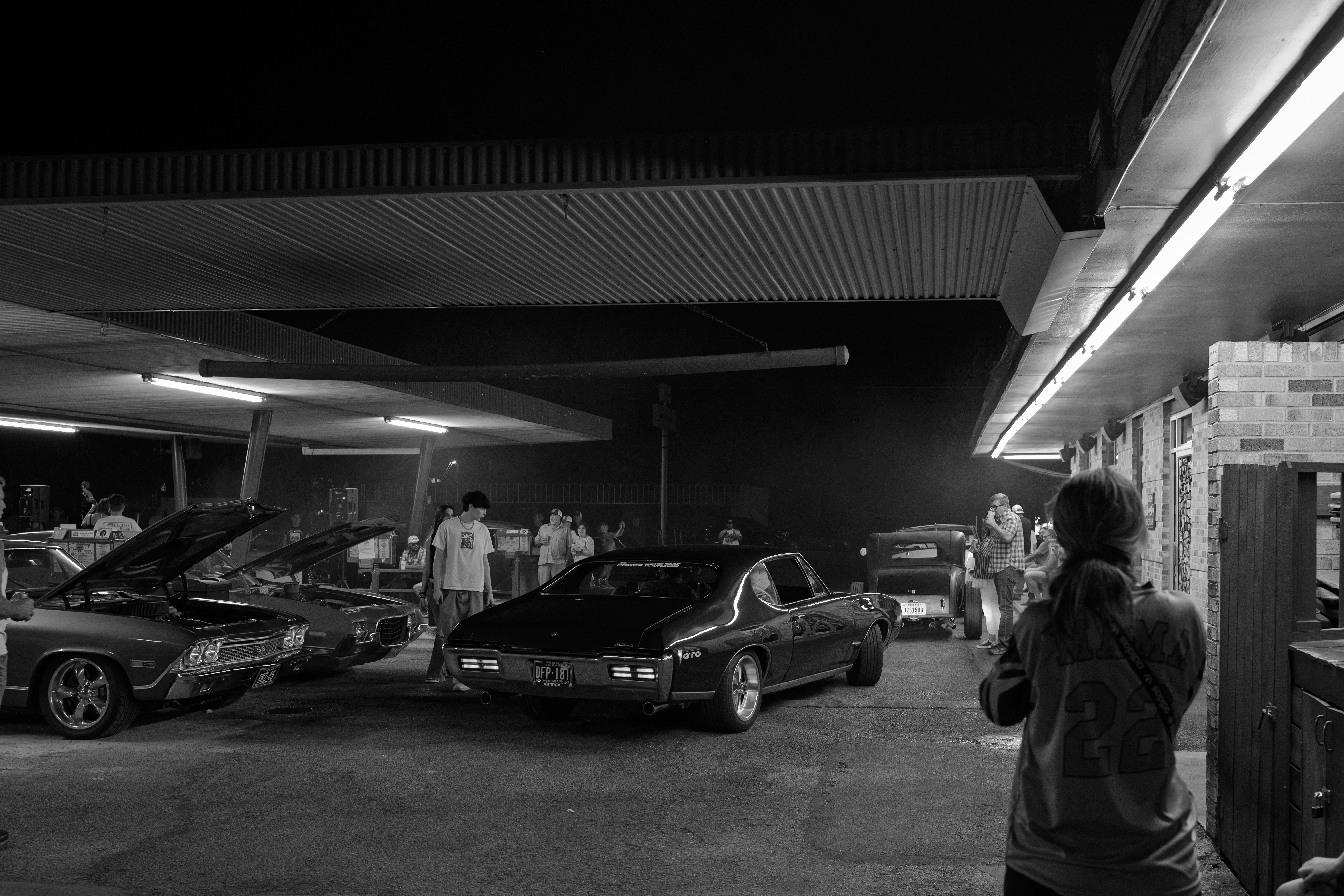 Black and white image of a nighttime car meet at a diner. Several classic cars with open hoods are parked, surrounded by groups of people conversing. Overhead fluorescent lights illuminate the scene. A person with a ponytail and a jersey stands in the foreground, capturing a photo. The atmosphere appears relaxed, with various people engaged in conversation or examining the cars.