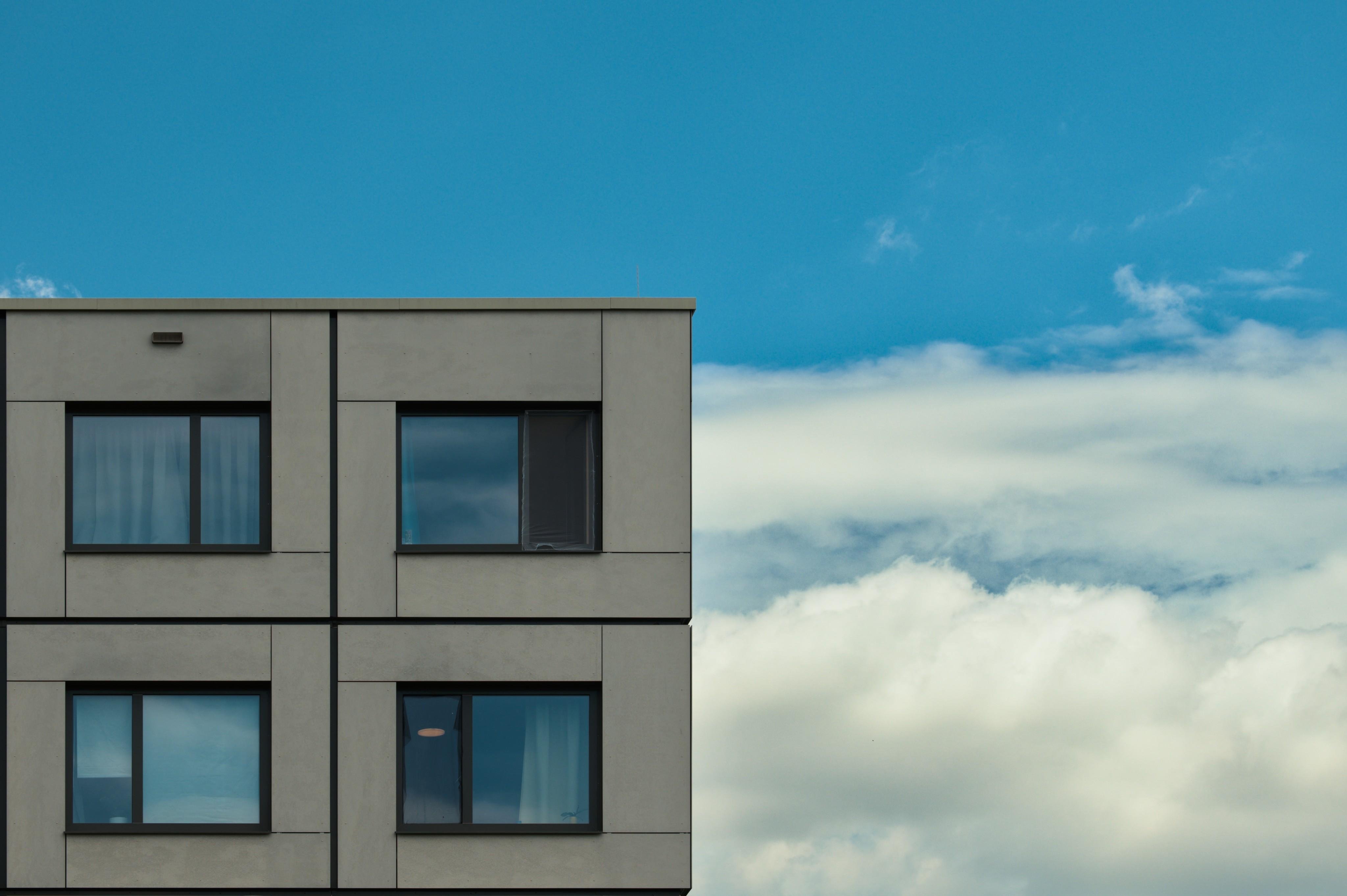 Right corner of a gray modern apartment building with four rectangular windows on the left, set against a blue sky with large white clouds on the right.