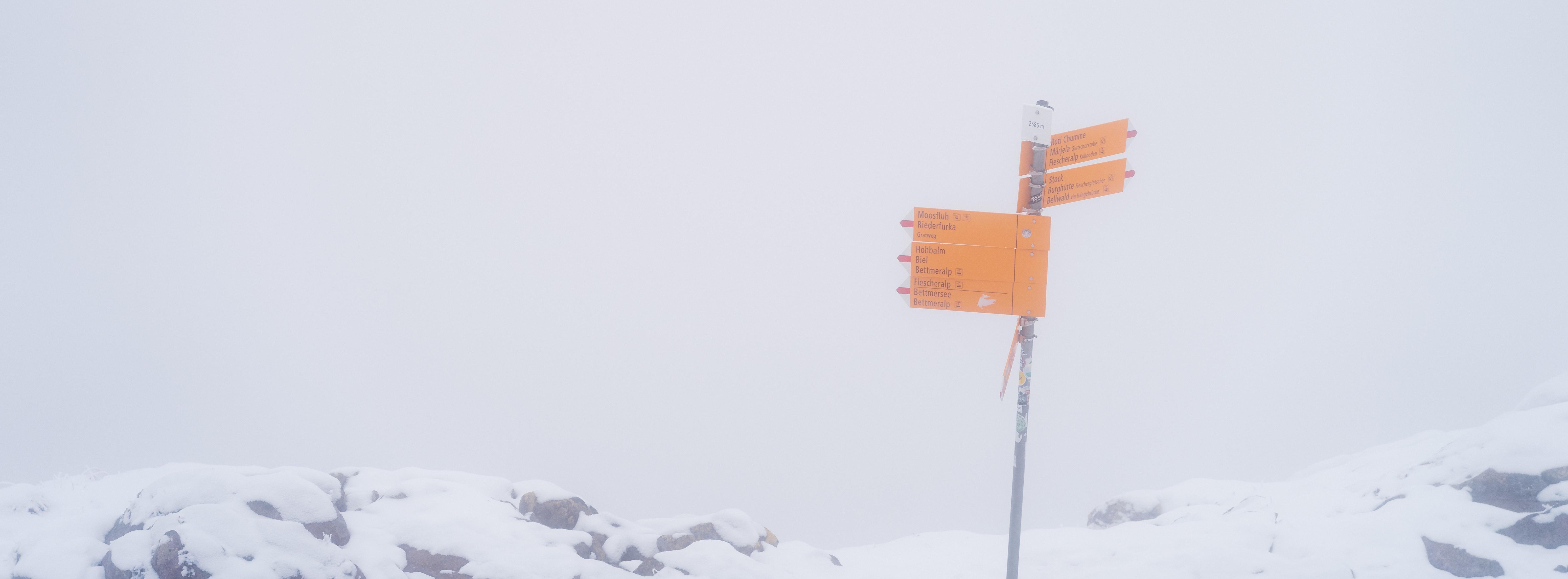 Orange signpost with multiple directional arrows stands amidst a snowy landscape; foggy background creates a muted atmosphere with snow-covered rocks visible at the base.