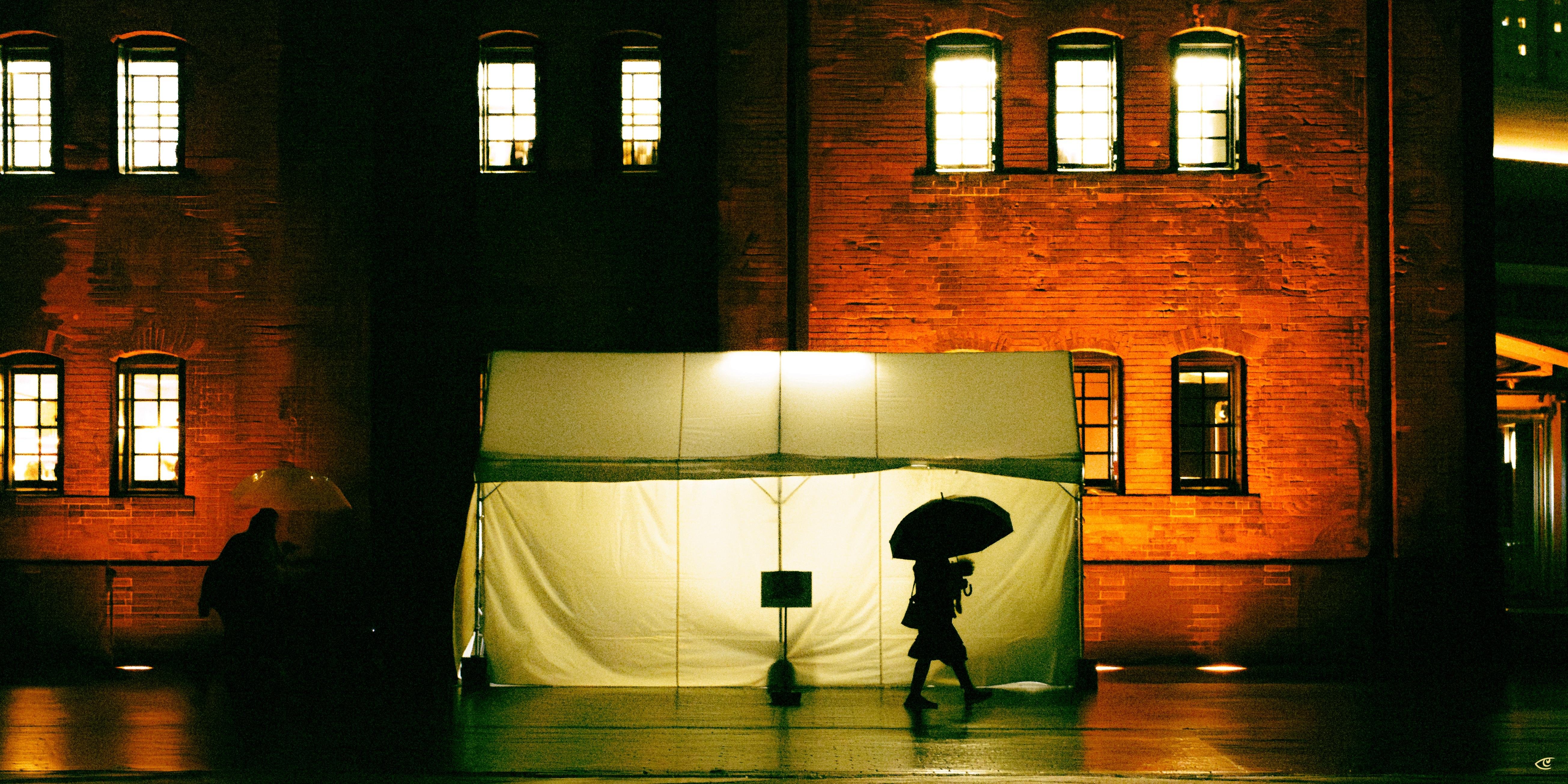 Nighttime street scene with a lit white tent in front of a red brick building, wet pavement reflecting light, and two silhouetted pedestrians holding umbrellas, one walking past the tent.
