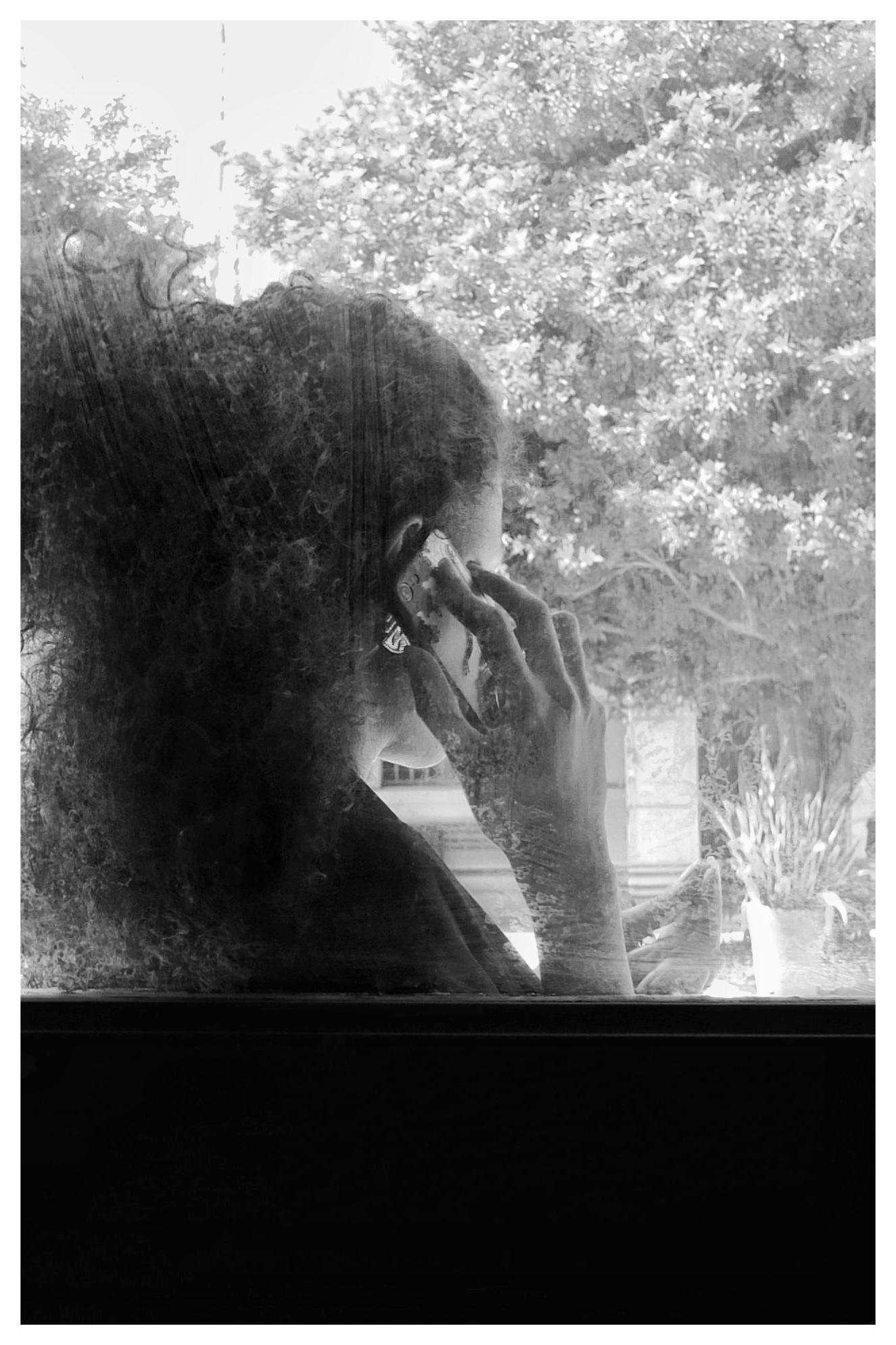 Side-profile view through a window of a person with curly hair holding a phone to their ear, with trees visible outside and a potted plant on the windowsill.