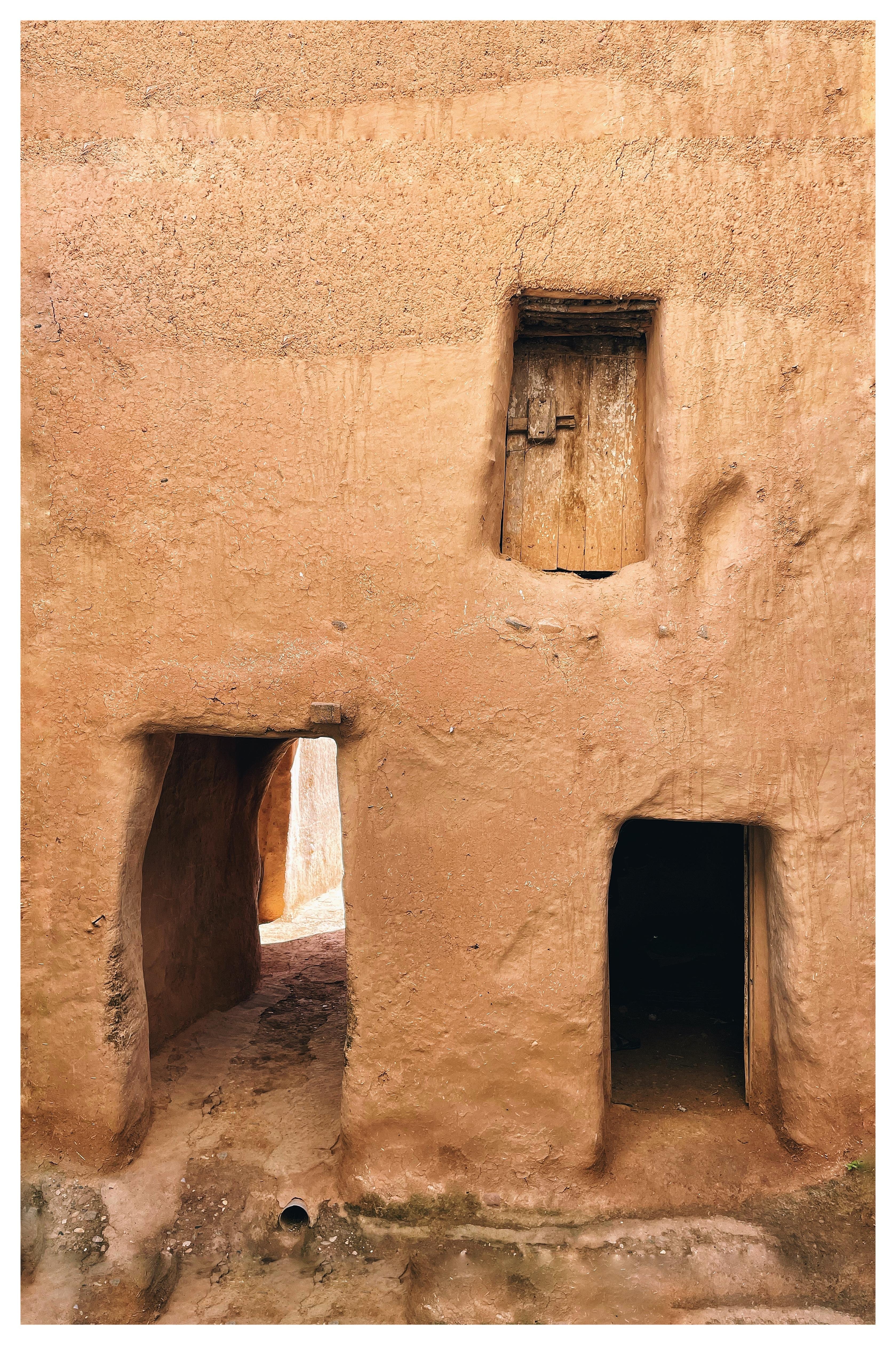 Tan mud-plastered wall with an arched passageway on the left, a dark rectangular doorway on the right, and a small recessed wooden door or shutter above; rough textured surface with cracks and worn edges.