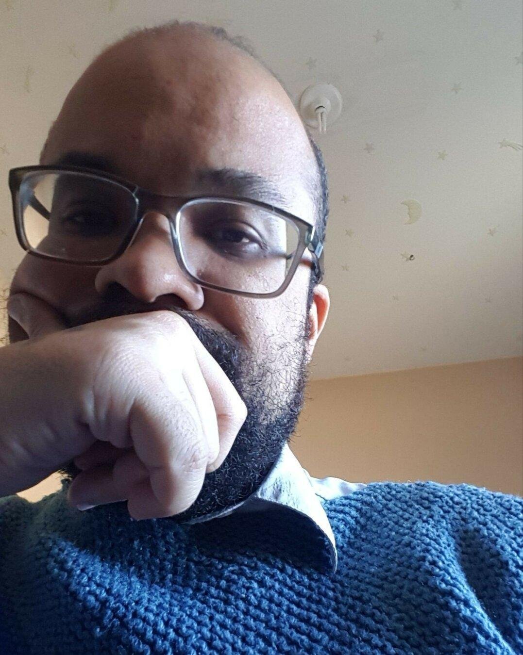 Man with glasses and a beard rests his chin on his hand. Wears a blue knit sweater over a collared shirt. Background features a light-colored ceiling with star and moon patterns.