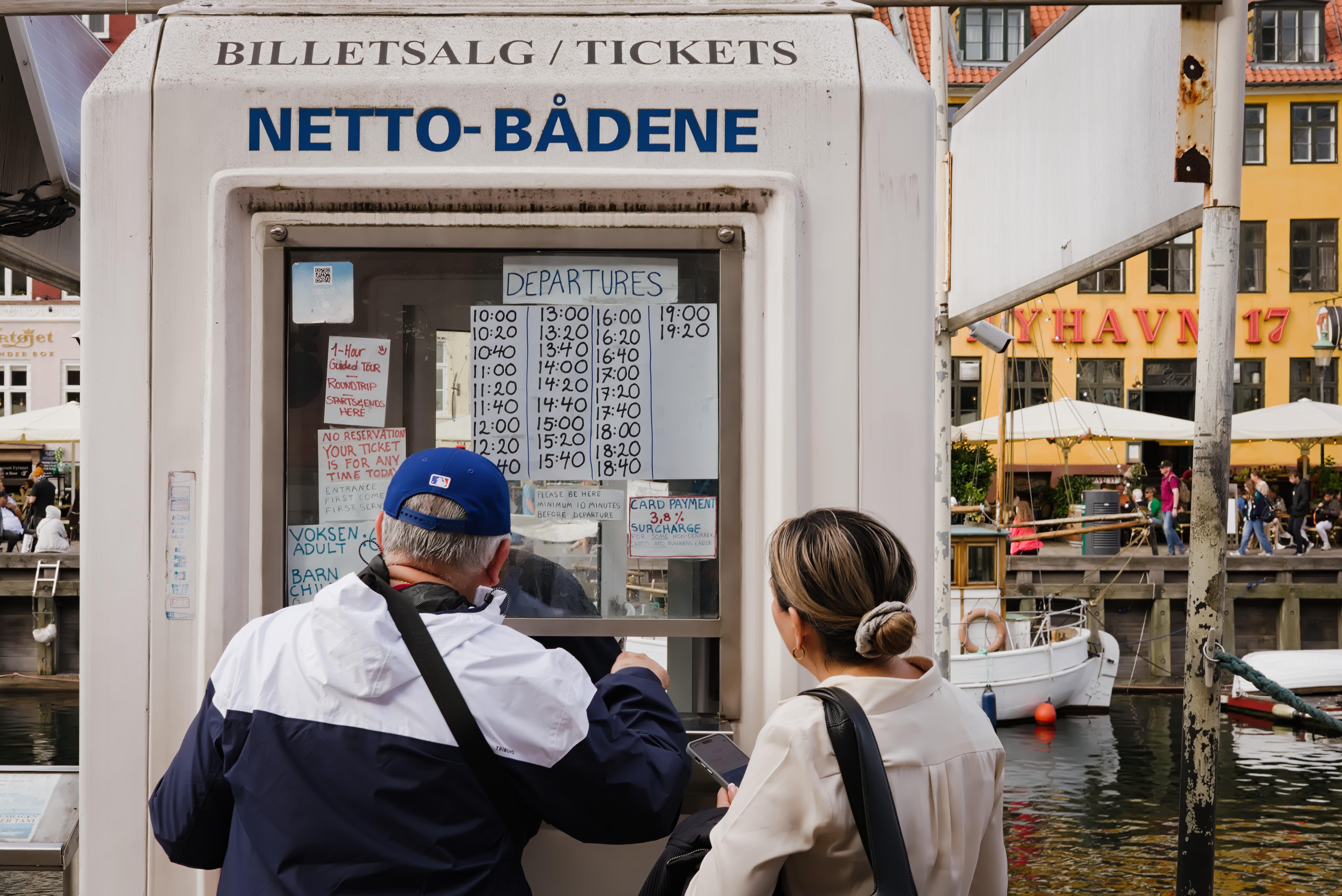 Two people stand in front of a white ticket booth labeled "Netto-Bådene" with a wide range of handwritten papers taped to the window. The man wears a blue cap and jacket, while the woman has a beige jacket and tied-back hair. In the background, colorful buildings and boats along a canal can be seen.