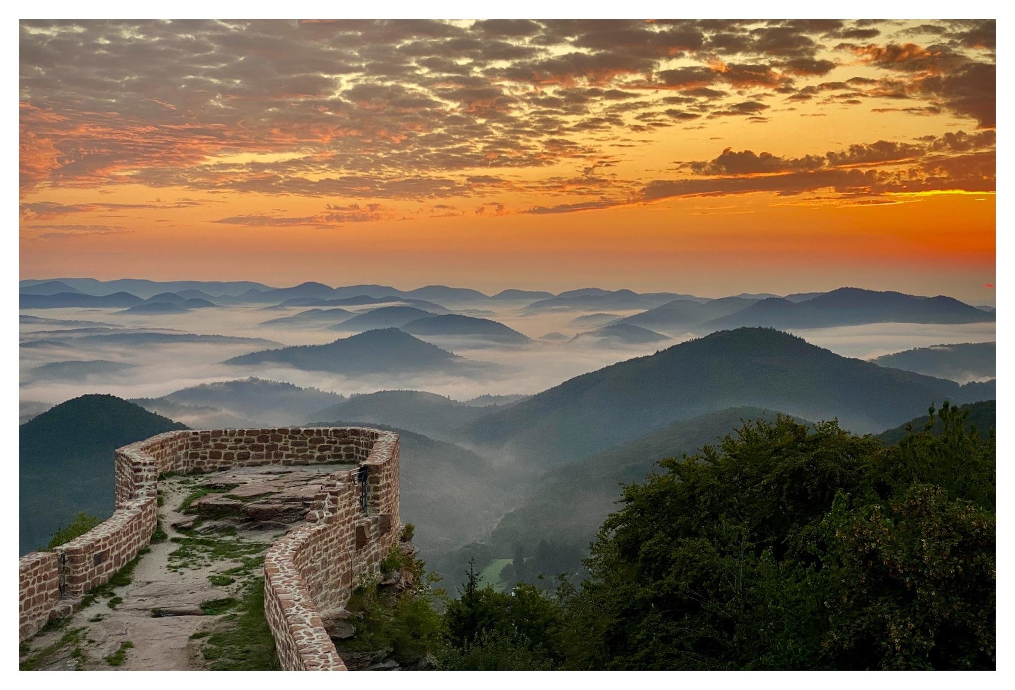 Curved stone lookout wall of the Wegelnburg foreground overlooking layered, misty mountain ridges, with a wide orange-and-gold sky filled with scattered clouds at sunrise.