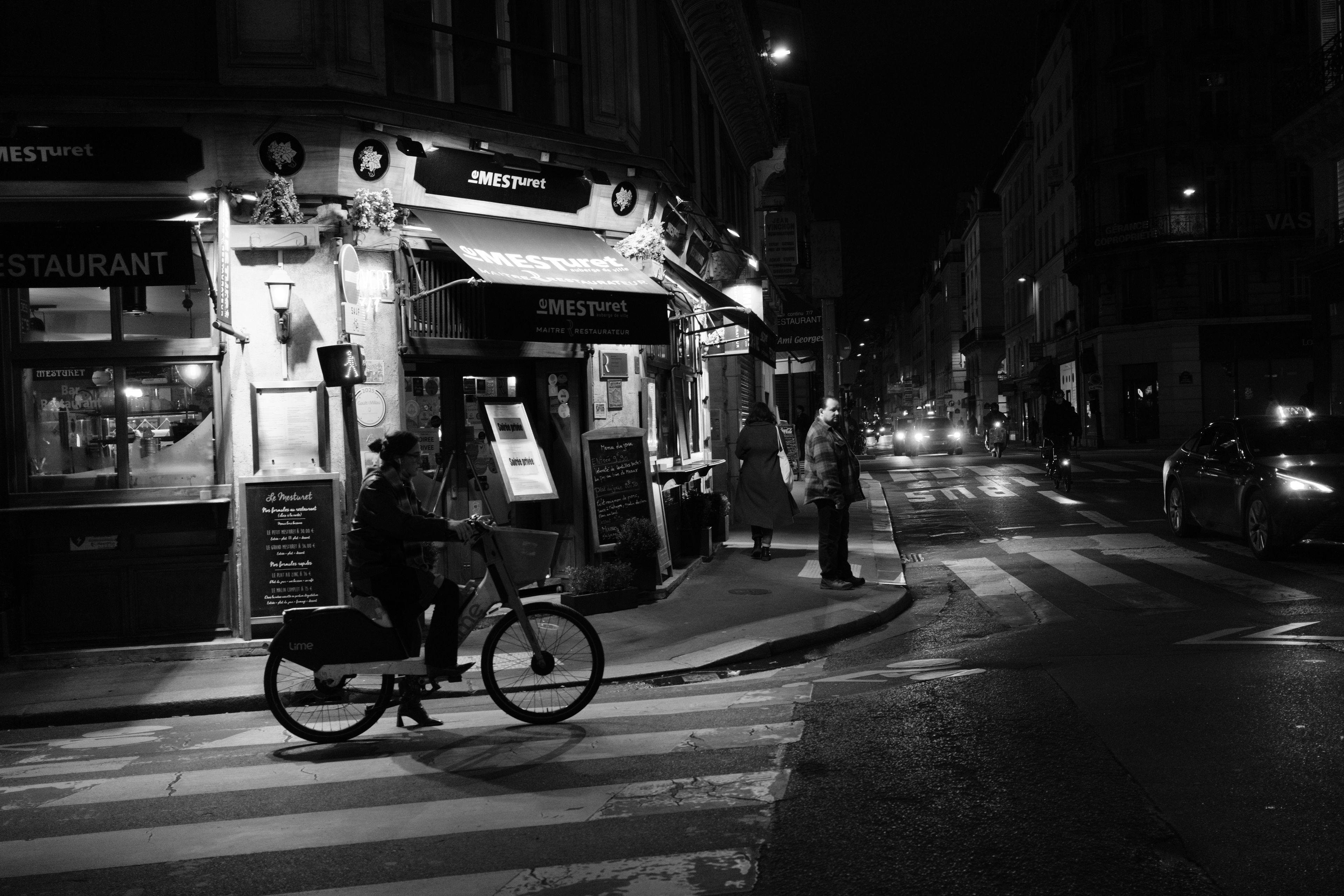Photo horizontale en noir et blanc. À un croisement, devant l’entrée d’un restaurant, une cycliste est arrêtée sur un passage piéton en attendant de pouvoir traverser le croisement. Un piéton attends aussi sur trottoir