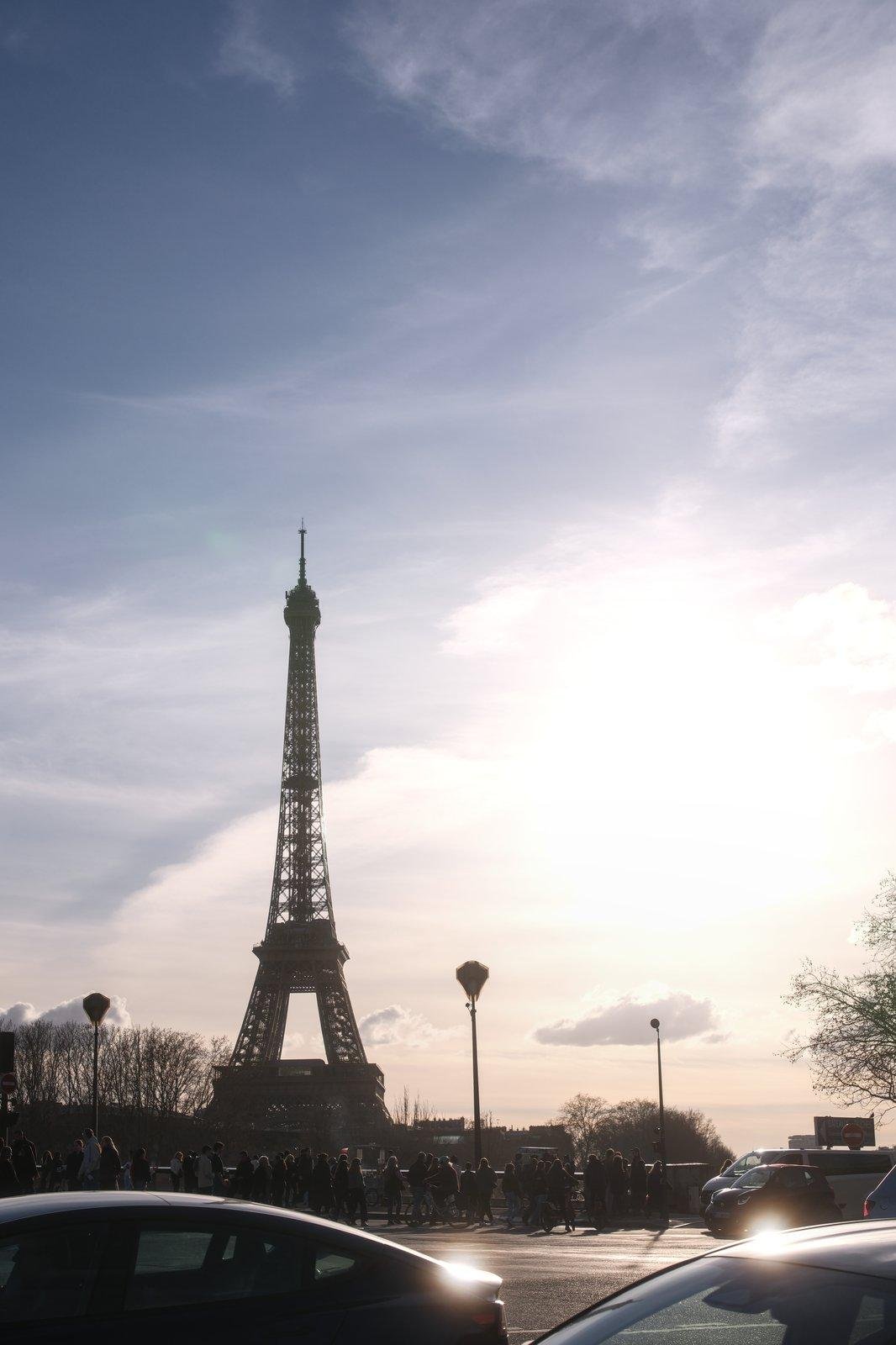 Photo verticale. La Tour Eiffel au loin, occupant environ 50% de la photo en hauteur, en contre jour une fin d’après midi de Février, le soleil arrivant de la droite de l’image. Le toit de 2 voiture reflèlent le soleil au premier plan, sous un ciel bleu avec des voiles de nuage