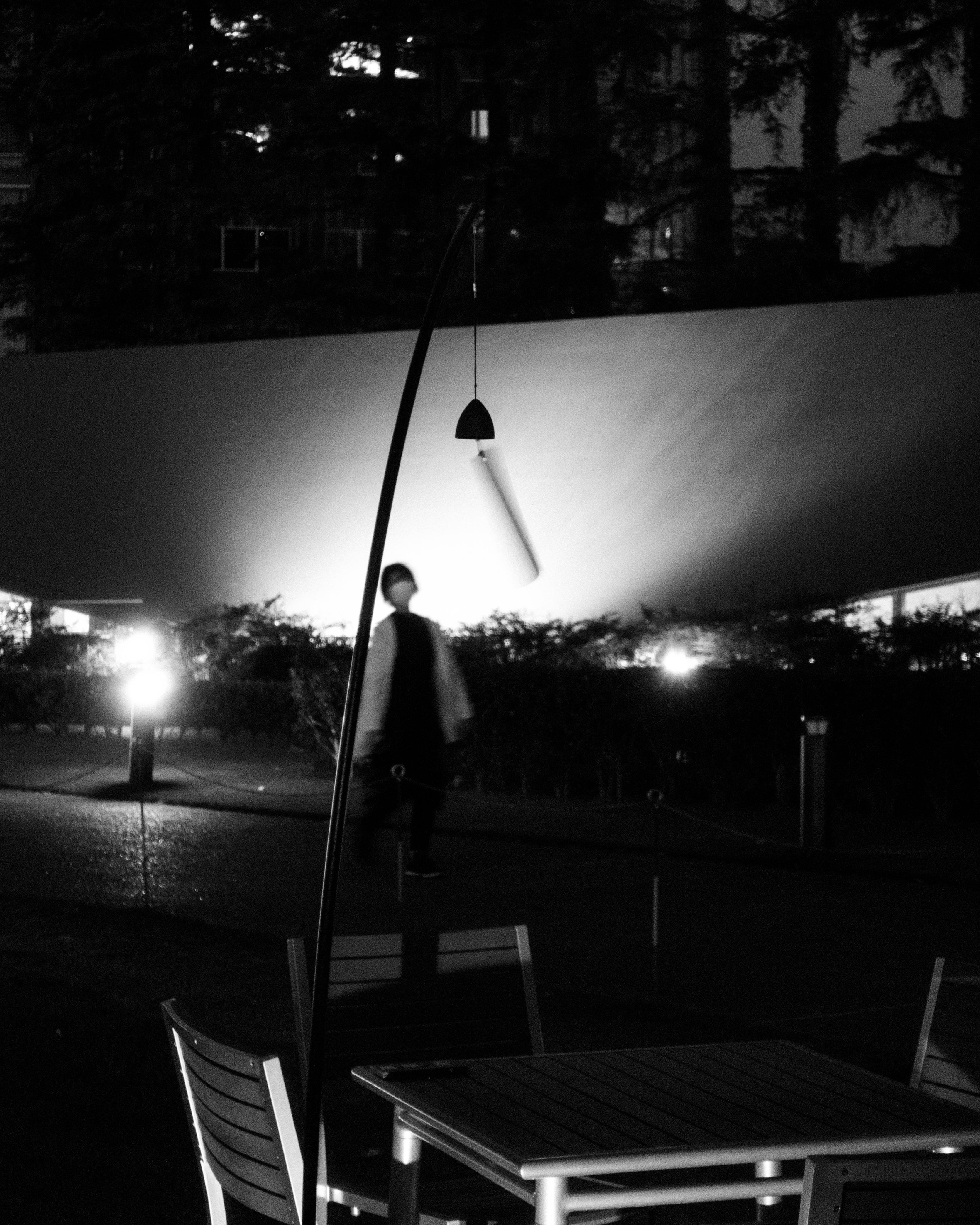 Monochrome outdoor scene with an empty table and chairs in the foreground. A modern, curved lamp post stands nearby. Background features a pedestrian walkway with a blurred figure walking by, illuminated by bright, diffused lights. Bushes and trees partially visible against a dark sky.