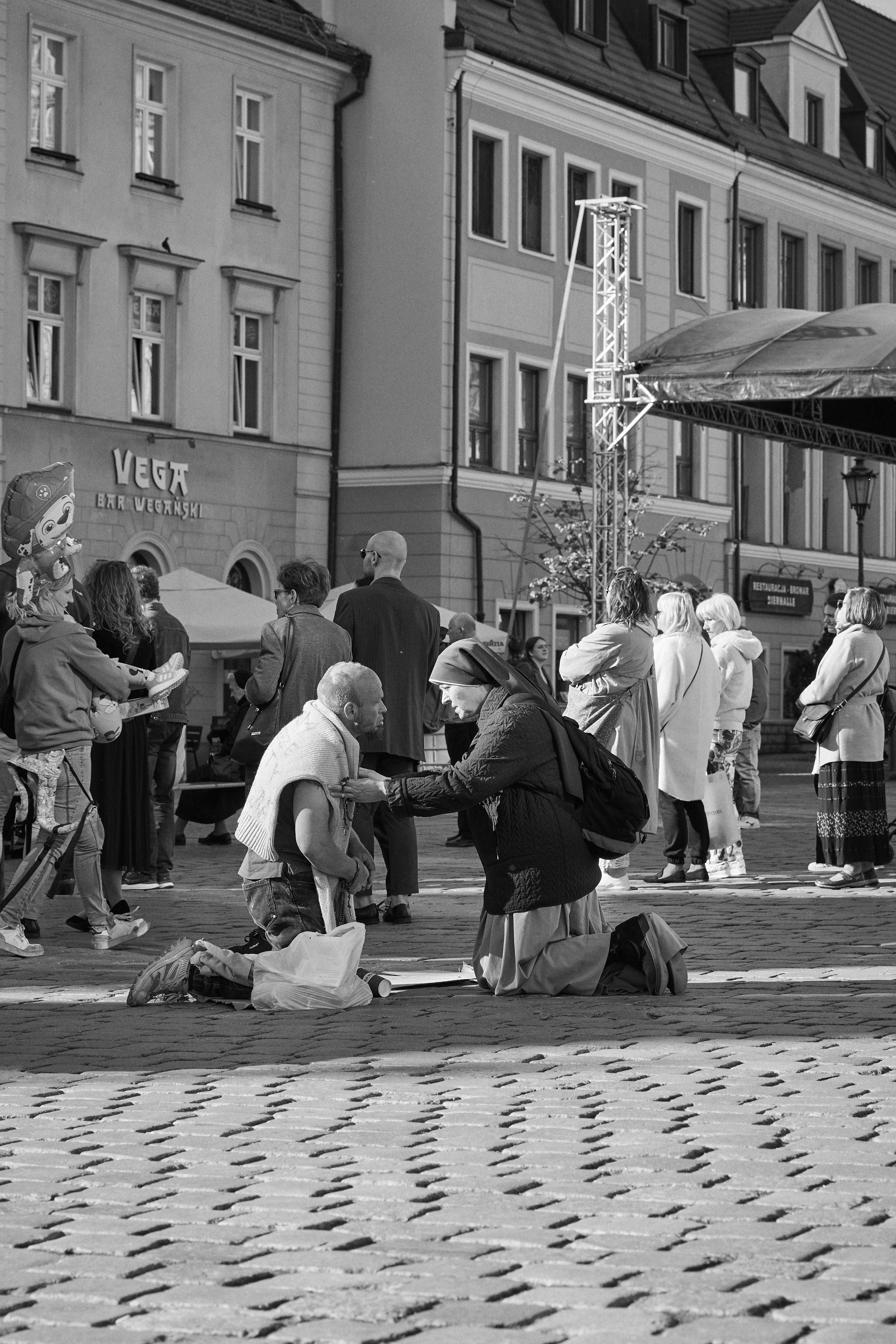 A black-and-white photograph depicting a street scene in which a nun is kneeling and engaged in conversation with a kneeling man on a cobblestone sidewalk. The scene is set in front of WrocΕawβs Town Hall. In the background, several individuals are visible standing and appearing to listen attentively.
