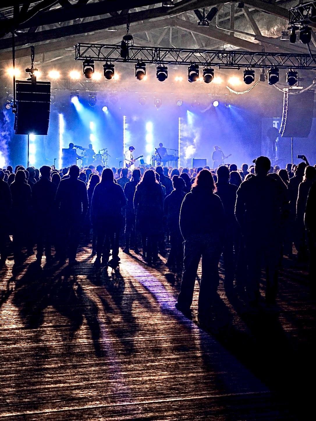 Silhouetted crowd standing on a wooden floor in an indoor concert venue, facing a stage with musicians, blue spotlights, haze, and vertical light bars; overhead truss with multiple stage lights and large hanging speakers.
