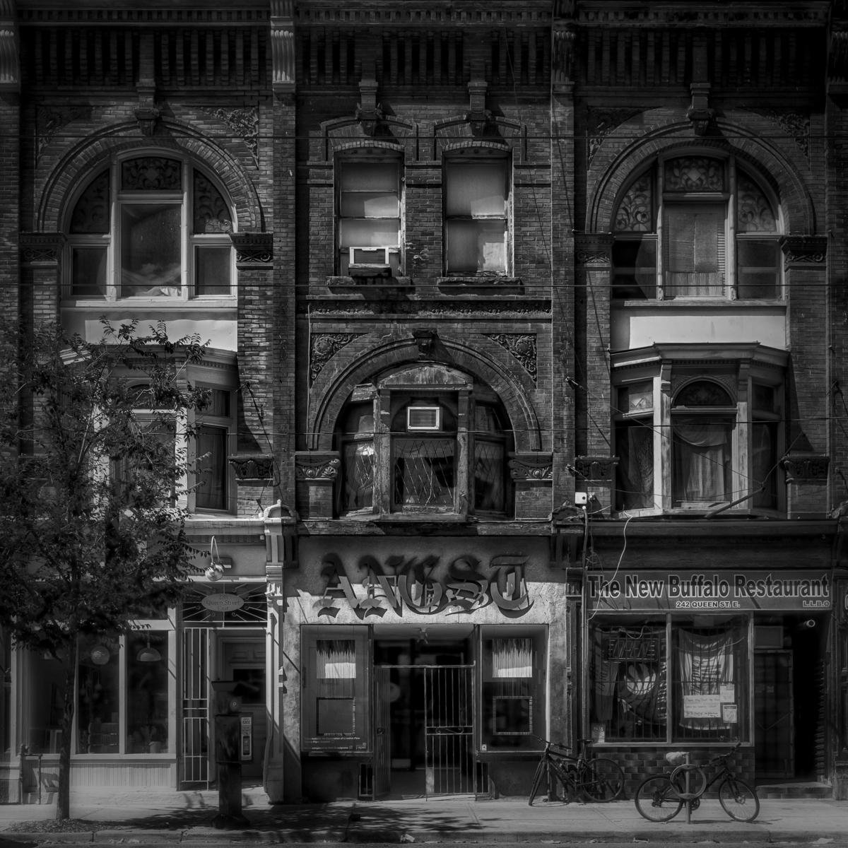 Facade of an old brick building with intricate architectural details, housing two businesses. The left side features a sign for "ANGST" with barred entrance, while the right side displays "The New Buffalo Restaurant" with large windows. A black bicycle leans against the wall near the restaurant. A tree is visible in the foreground, casting shadows on the sidewalk.