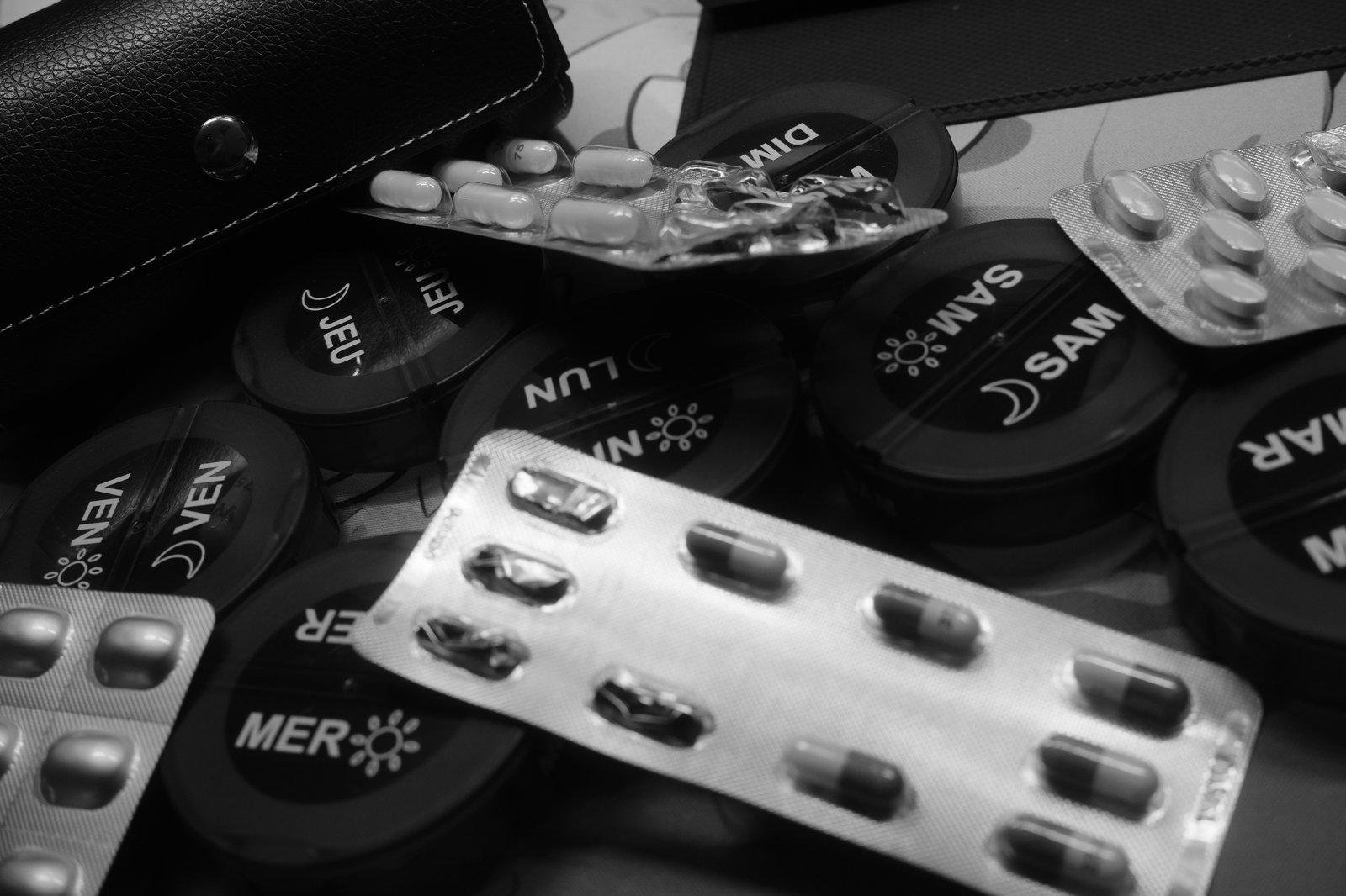 Photo horizontale en noir et blanc. Montrant des boîtes d’un pillulier en forme de palet (un par jour) et des plaquettes de médicaments en vrac dessus