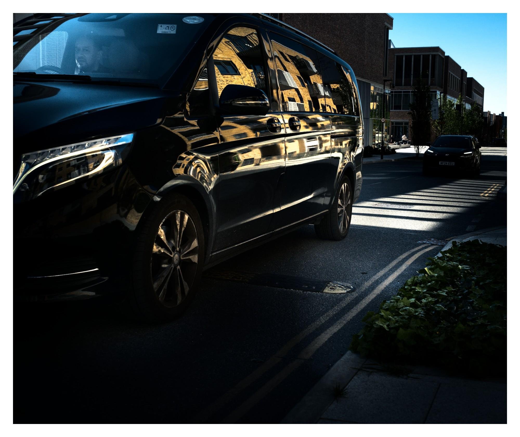 Black van in the left foreground on a city street, its side reflecting nearby buildings; another car drives in the background near a pedestrian crossing with long shadows, with modern brick and glass buildings and a sidewalk with plants on the right.
