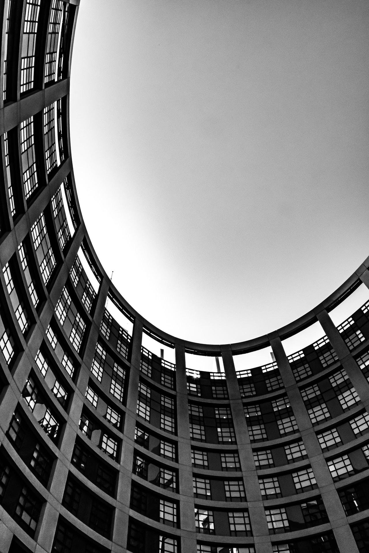 Curved facade of a modern building with multiple floors featuring rows of square windows. The shot is taken from below, looking up at the clear sky framed by the building's architectural curve. Black and white filter emphasizes the structure's geometric pattern and contrast.